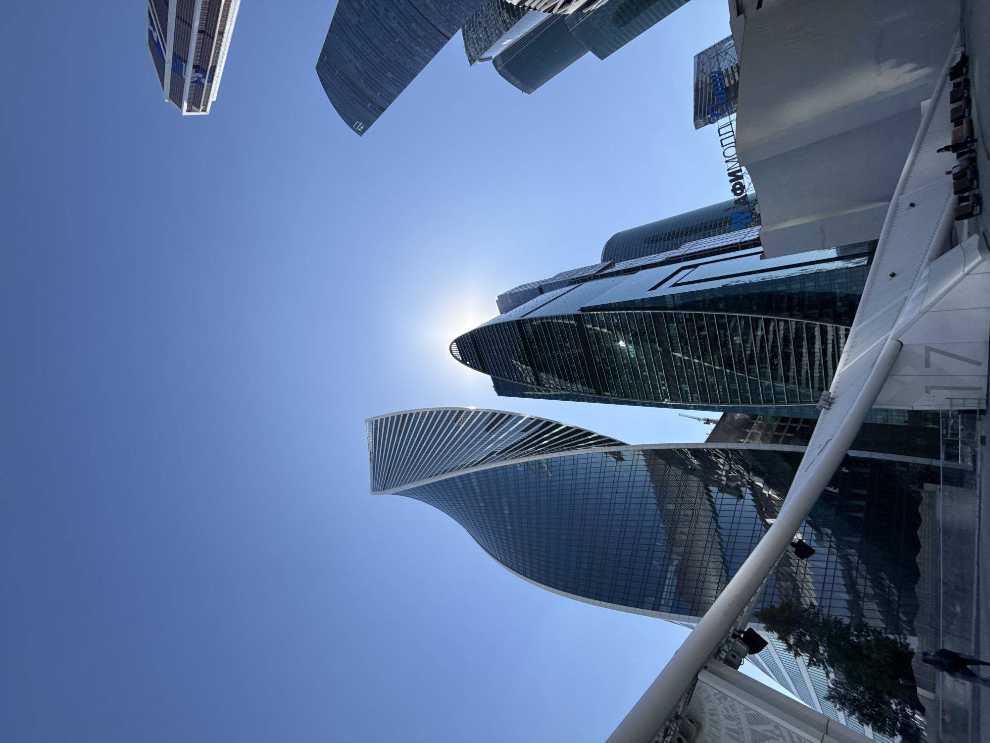 This is a low-angle shot depicting the modern high-rise architecture of the Moscow International Business Center (Moscow-City) in Moscow, Russia, under a clear, bright blue sky. The sun is positioned high, creating a strong sunburst effect directly behind one of the central skyscrapers, causing significant backlighting.
In the left foreground, the distinctive spiraling form of the Evolution Tower dominates, its glass facade reflecting the sky. To its right, another prominent skyscraper is partially obscured by the intense sun glare. Multiple other glass-clad skyscrapers of varying heights and designs are visible across the frame, receding into the background on both the left and right sides. On the far upper right, a portion of a curved skyscraper displays a corporate logo or insignia.
A large, white, geometrically patterned pedestrian structure, possibly part of a bridge or walkway, extends diagonally from the lower left to the middle right of the image. The number "7" is visible on one of its white panels. In the lower right, the "АФИМОЛЛ Сити" (AFIMALL City) sign in blue Cyrillic letters is mounted on a white, angular building that forms part of the complex.
Several individuals are present in the lower portion of the scene: one person walks leftward near the base of the white structure in the lower left, and two individuals are seated on benches near the "AFIMALL City" building in the lower right. Reflections of other buildings and the sky are prominent on the glass surfaces of the skyscrapers. A small green tree is visible behind the white structure in the lower left, and a construction crane can be faintly observed between buildings in the mid-ground.