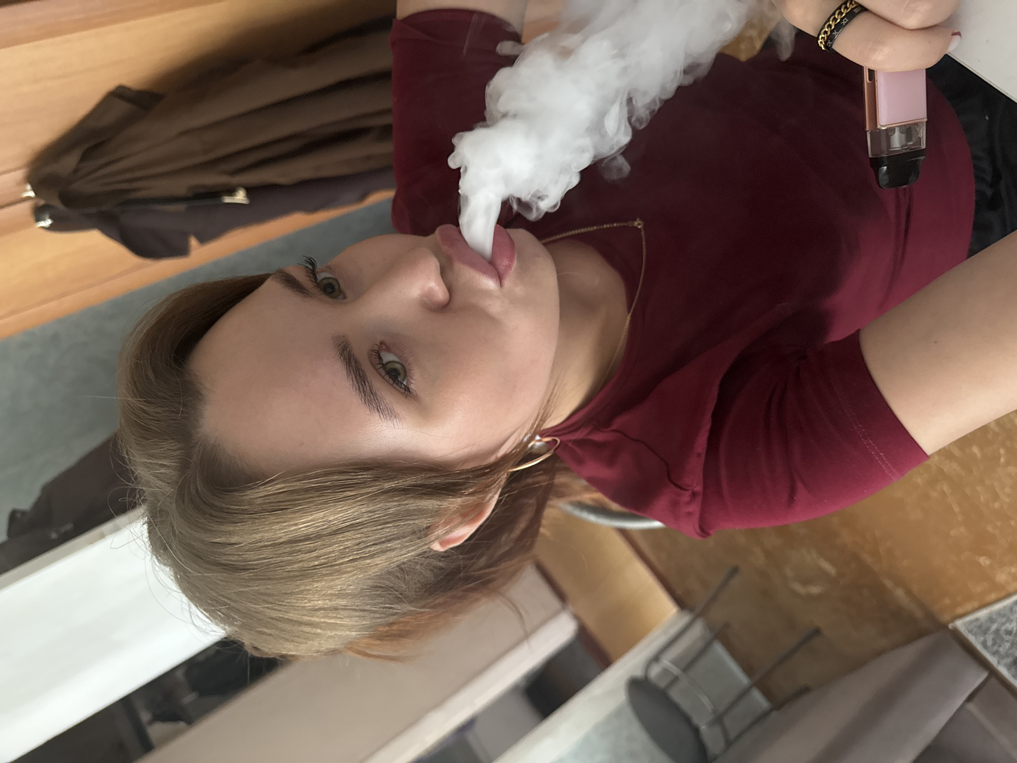 In this media file, a young person is captured exhaling a plume of white vapor, likely from an electronic cigarette. The individual has light skin and light brown hair, which is styled loosely. They are wearing a maroon-colored shirt and a gold necklace. A pink and rose gold colored electronic device is visible in their right hand, held near their chest. The person also wears a ring with a gold chain pattern on their right index finger and a large hoop earring on their left ear. Their eyes are open and looking towards the camera, and their tongue is slightly extended, interacting with the vapor. The background suggests an indoor setting with wooden furniture and a tiled floor. The lighting appears to be artificial, indicating it could be daytime or nighttime. There is no visible text in the image. The scene appears to be casual and personal.