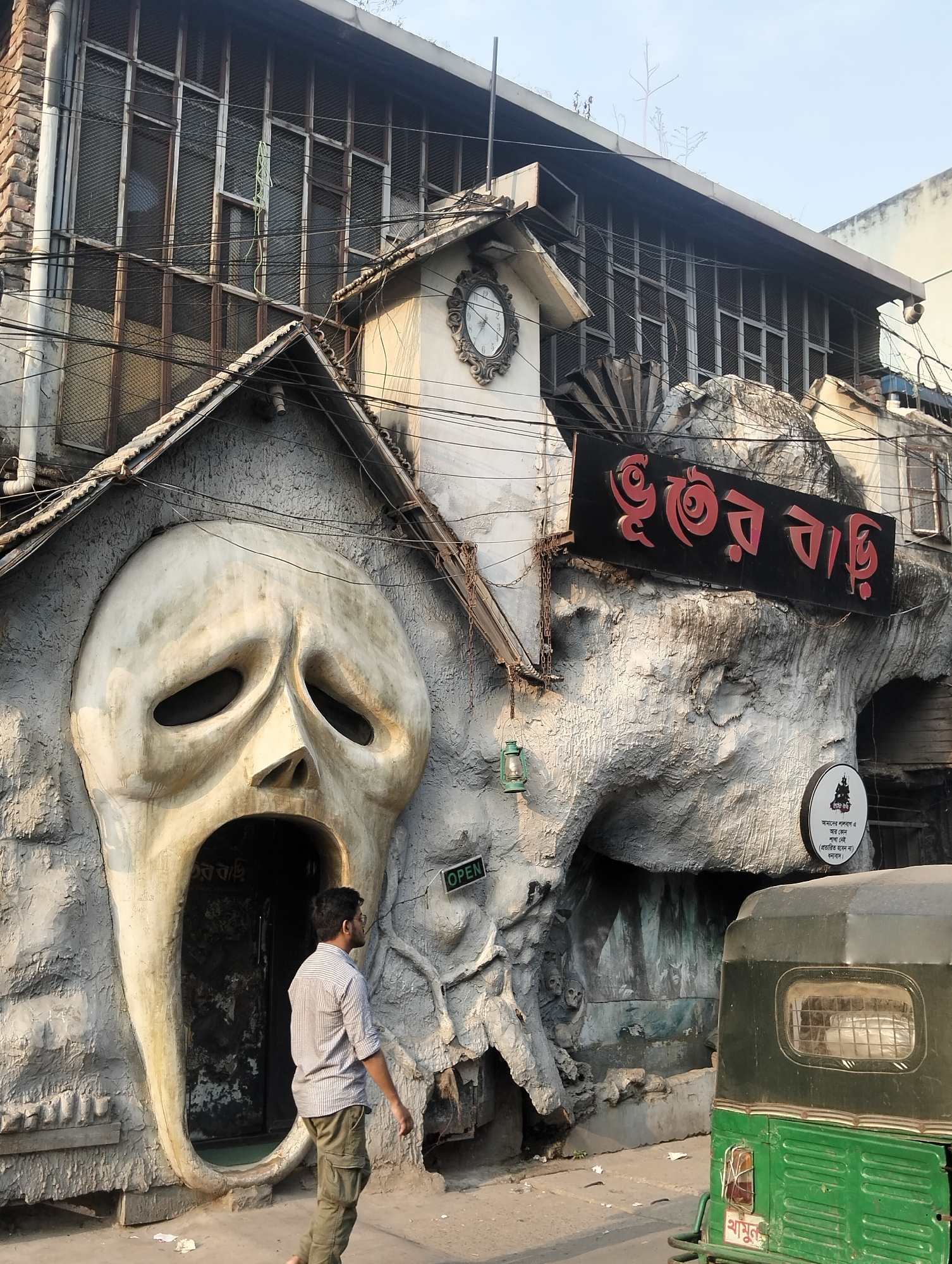 The image shows the Bhuter Bari (Ghost House) restaurant in Dhaka, Bangladesh. A man wearing a light-colored, striped, short-sleeved shirt and khaki pants is walking on the sidewalk in front of the restaurant. The building is constructed to look like a haunted house with a large, stylized screaming face forming the entrance to one section. Another section of the building is built to resemble a cave. There is a sign in Bengali that reads "Bhuter Bari" above the cave entrance. A sign reading “OPEN” is visible next to the screaming face. A green auto rickshaw (CNG) is parked on the street in front of the building, partially obscuring the cave section. The upper part of the building has windows with metal mesh. Power lines crisscross the scene. The sky is light.