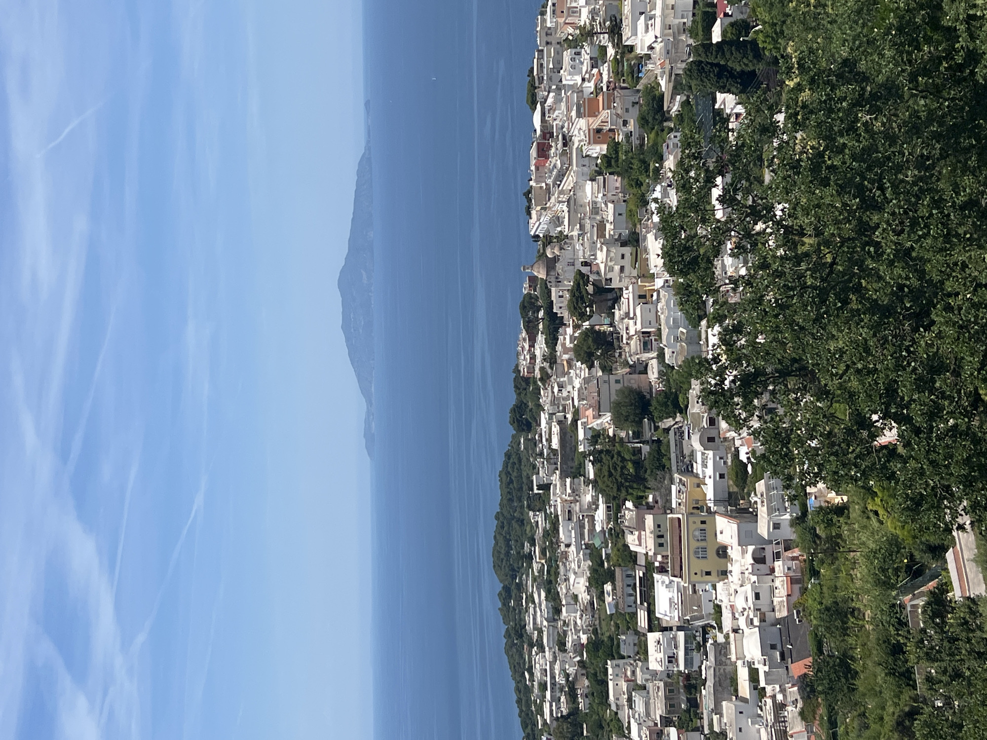 The image is an aerial view of Anacapri, Italy, a town on the island of Capri in the Bay of Naples. The scene is dominated by the vibrant blue of the Tyrrhenian Sea in the background, with a distant island visible on the horizon. In the foreground, the town of Anacapri unfolds, characterized by its white-washed buildings with terracotta roofs, clustered together on a hillside. Lush green trees and vegetation are interspersed throughout the town and surround it, suggesting a Mediterranean landscape. The sky is a clear blue with scattered white clouds, indicating a sunny day. The image is a static, still shot, capturing a panoramic view of the coastal town and its surrounding environment.