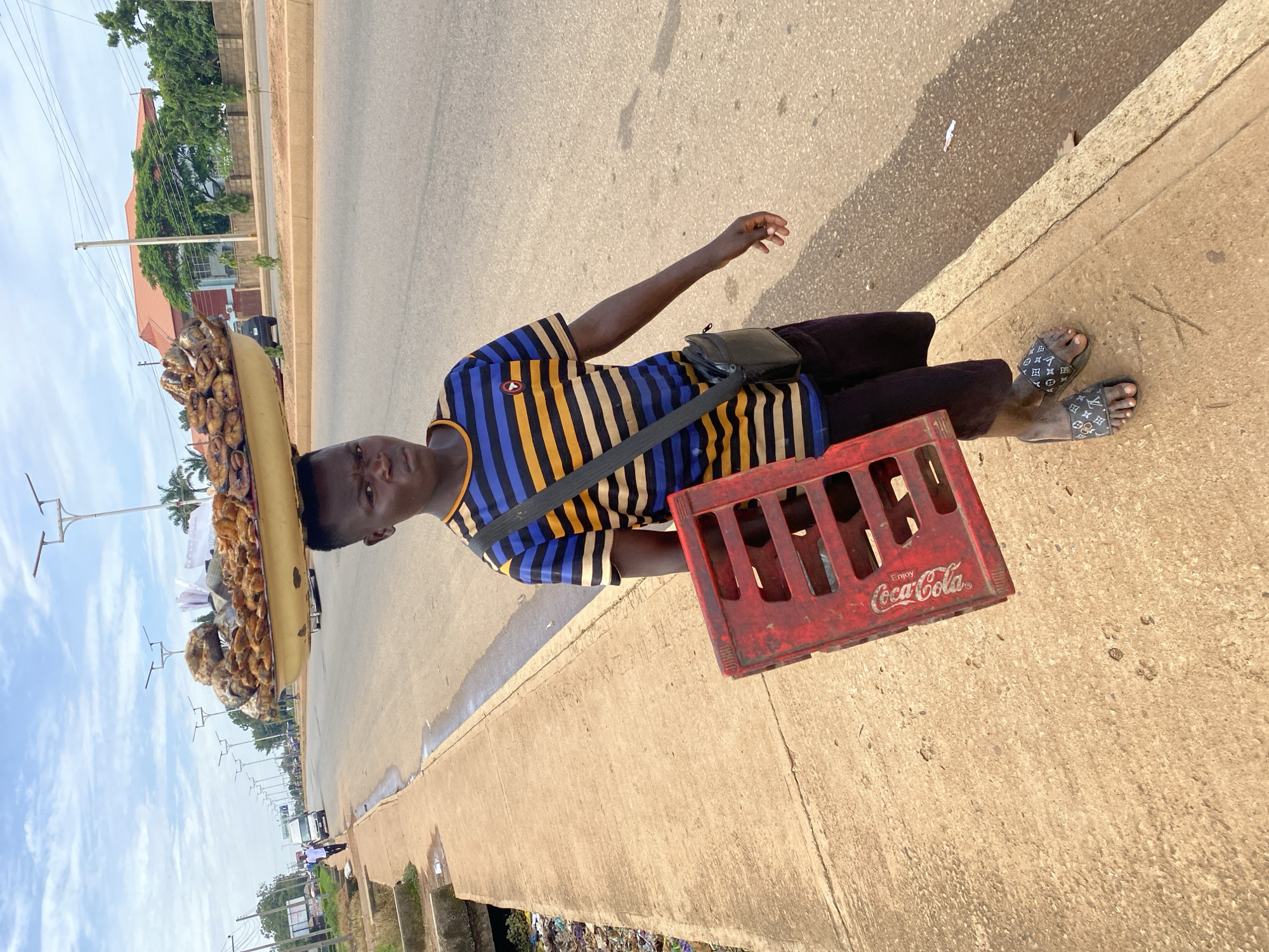 A young Black male vendor is walking on the side of a paved road in Bukuru, Nigeria. He is balancing a large, round tray filled with fried fish on his head. He is wearing a horizontally striped t-shirt with blue, yellow, and black stripes, dark colored shorts, and sandals with a designer pattern. He is carrying a red Coca-Cola crate in his left hand. A black shoulder bag is slung across his chest. The road is flanked by a concrete sidewalk and a drainage ditch filled with debris. In the background, there are buildings, trees, streetlights, and passing vehicles. The sky is partly cloudy.