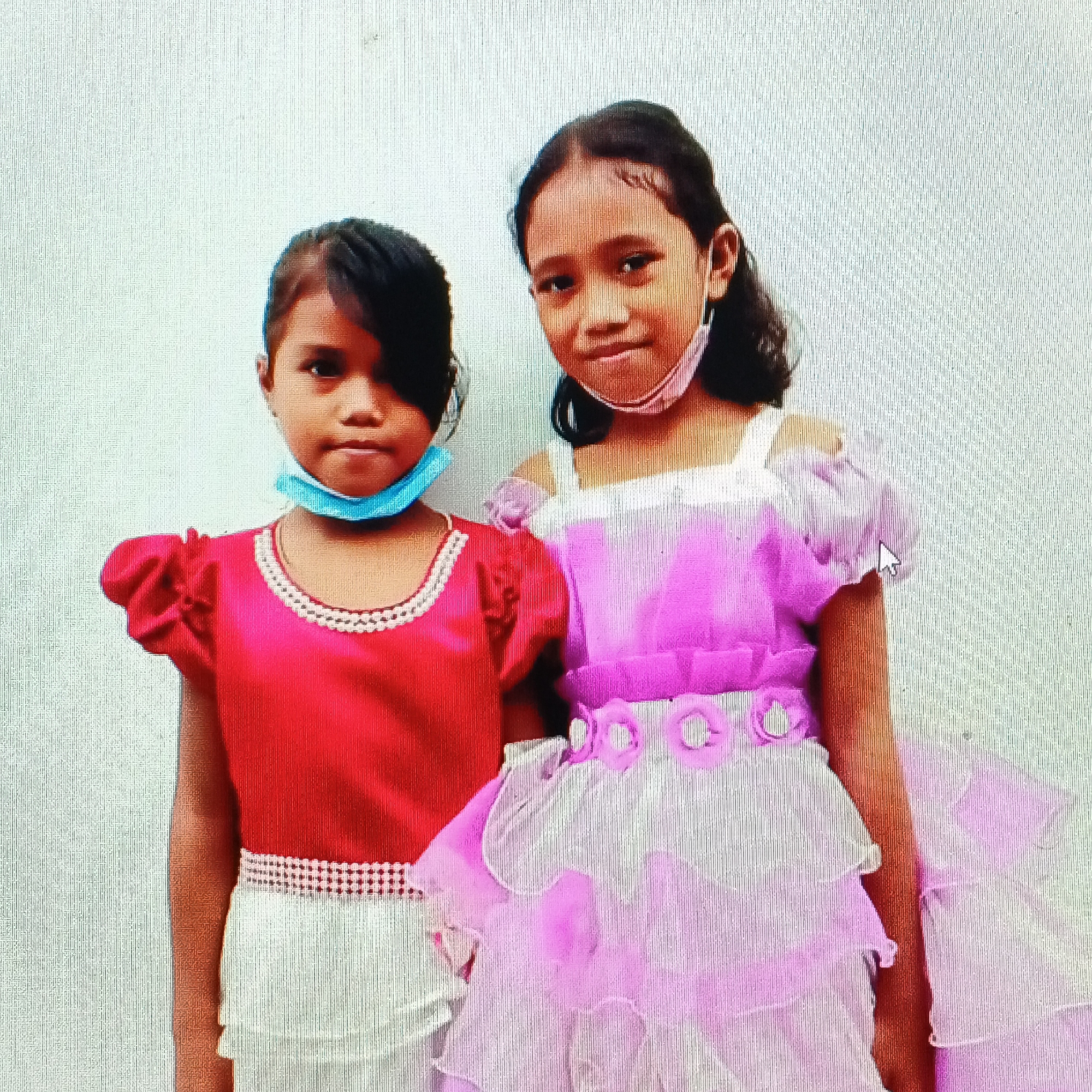 Two young girls, one wearing a red top with a white-trimmed neckline and a white skirt, the other in a light pink dress with a ruffled hem and sleeves, stand side-by-side against a light-colored, textured wall. Both girls have dark hair and are wearing face masks. The girl on the left has a light blue mask, and the girl on the right has a light pink mask. The girl on the left is wearing her mask below her nose, while the girl on the right has hers positioned correctly. The girl on the right's dress features floral embellishments around the waist and hem. The image appears to have been taken indoors or in a covered outdoor area, with a white or light gray background that has a faint vertical line texture.