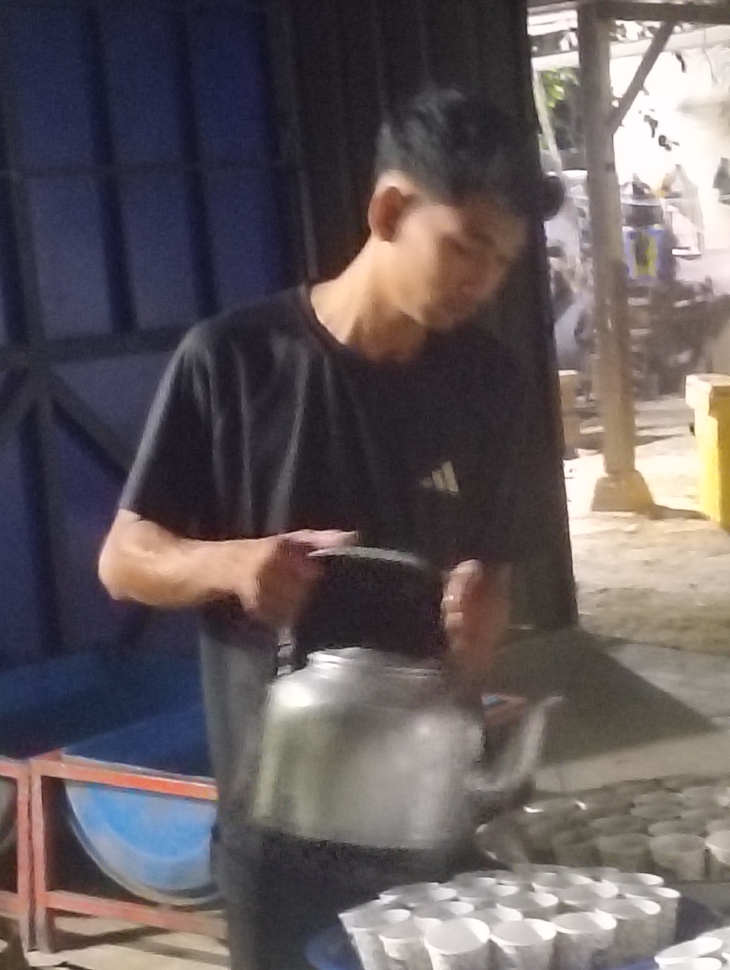 In Teungoh, Indonesia, a young male wearing a black t-shirt is captured in a dimly lit outdoor setting. He is in the process of pouring liquid from a metal kettle into a row of small, white paper cups arranged on a table. The background is blurred, but suggests an open-air market or street stall environment with various structures and possibly other individuals out of focus. The lighting is low, creating a somewhat grainy image.