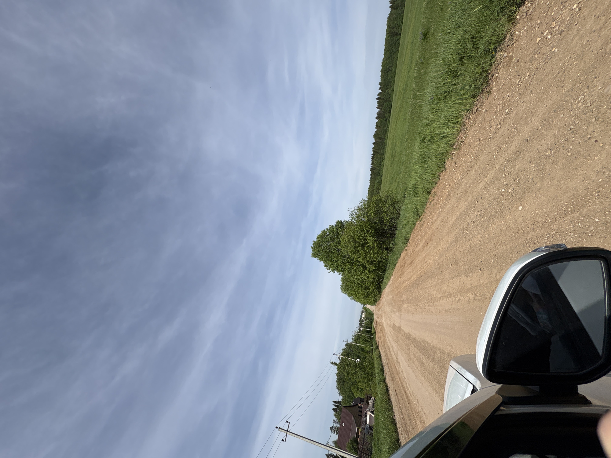 The image shows a view from a vehicle traveling on a dirt road. The road is flanked by grassy areas and trees on one side, and a field on the other. Power lines and houses are visible in the distance. The sky is overcast with scattered clouds. The rearview mirror of the vehicle is visible in the lower right corner of the frame. The location is Khokhlovka, Russia.
