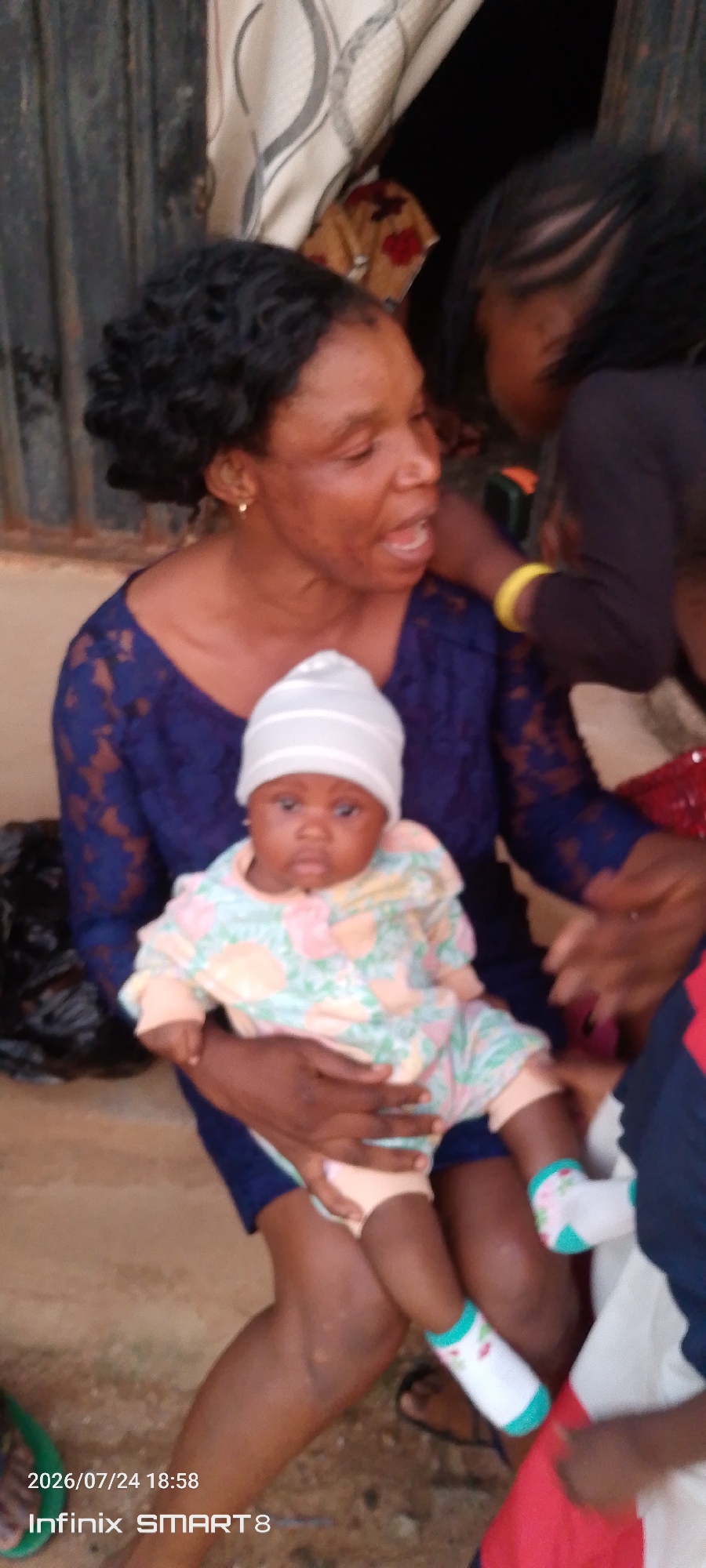 In Yelwa, Nigeria, a woman in a blue lace dress holds a baby wearing a white cap and a floral onesie. Another child, seen from behind, leans in towards the woman. The background is a blur of outdoor elements, possibly a residential area with a corrugated metal fence and some fabric hanging. The overall scene suggests a moment of familial interaction and care.