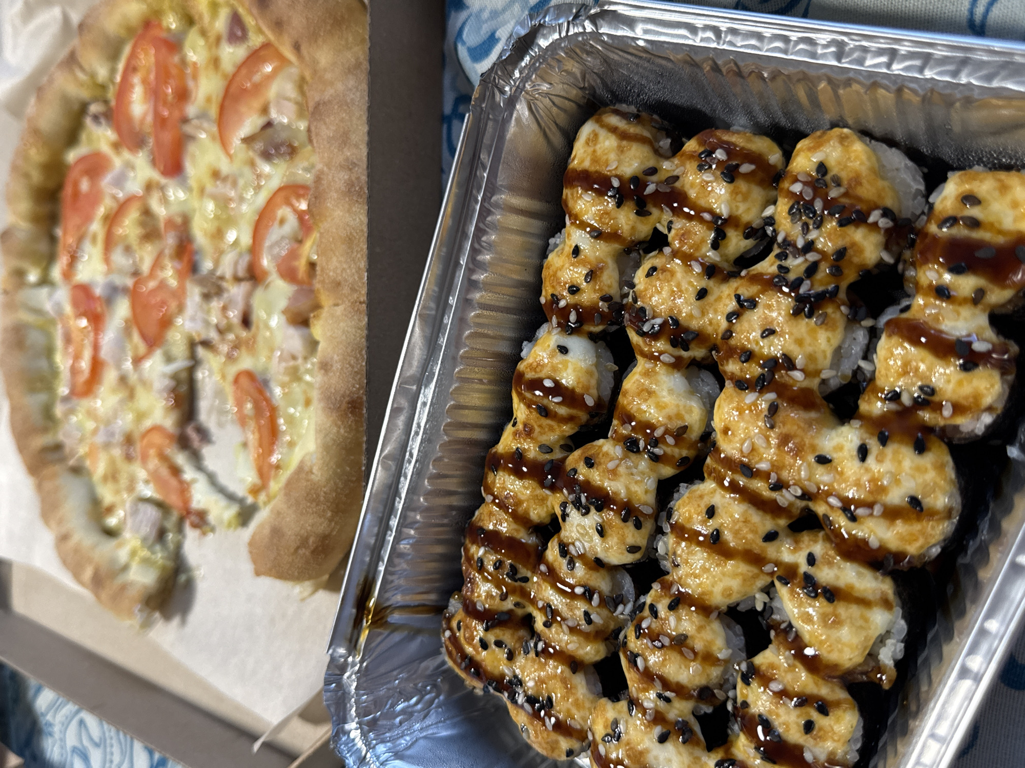 A close-up, high-angle shot shows a foil container of baked sushi rolls topped with a creamy sauce, sesame seeds, and a drizzle of dark sauce, positioned in front of a partially eaten pizza in a cardboard box. The pizza has a thick crust and is topped with melted cheese, tomato slices, and pieces of meat. The scene appears to be indoors, possibly on a table, with a patterned tablecloth visible underneath. There are no people or animals in the frame, and the focus is on the food. The location of Яромінскі сельскі Савет, Belarus is not depicted in this image.