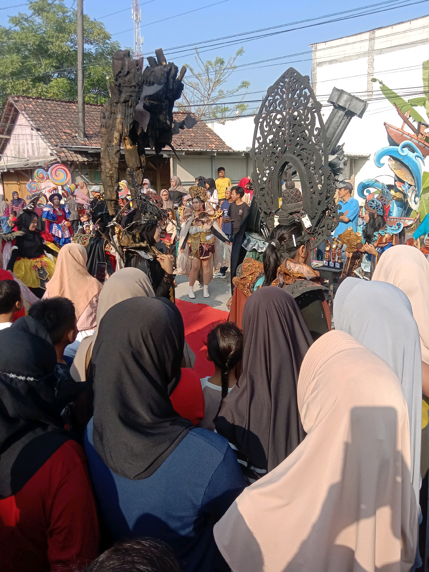 The image captures a street event likely in Sambongbangi, Indonesia, with a clear blue sky and a bright, sunny day. In the foreground, a crowd of people, many wearing hijabs, are gathered, with their backs to the camera, observing an event. Some children are also visible in the crowd. In the midground, participants in elaborate costumes are present. One individual is dressed in a dark, intricate outfit, possibly a costume depicting a dark entity or mythical creature, with a headdress adorned with branches and dark fabric. Another person is in a lighter, more ornate costume, possibly representing a royal or mythical figure, with gold accents and flowing fabrics.
Behind the costumed individuals, there are various decorative elements and structures. A large, dark, ornamental archway with intricate carvings stands prominently. To the right, there's a structure resembling a ship or boat integrated with wave-like blue and white decorations, featuring a fish motif. In the background, buildings with tiled roofs are visible, along with trees and utility poles, suggesting an urban or semi-urban environment. A radio tower can be seen in the distance. The overall scene depicts a festive atmosphere, possibly a parade, festival, or cultural performance.