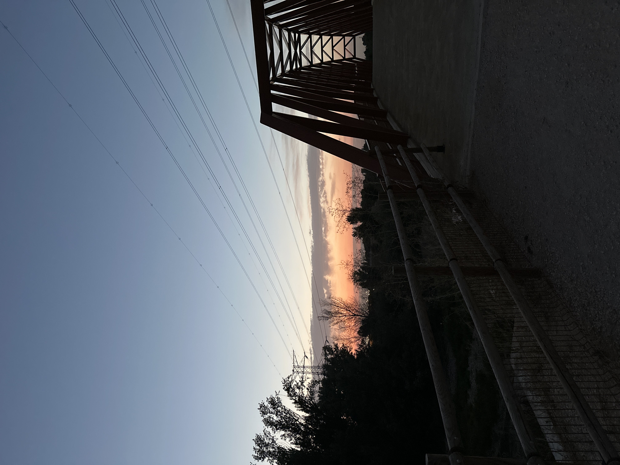 The image captures a sunset scene in Mejorada del Campo, Spain, featuring a pedestrian bridge spanning a dry creek bed. The bridge, constructed with dark red metal trusses, is the prominent subject in the foreground, with its walkway leading into the scene. To the left, silhouetted trees and foliage line the bank of the dry creek, with a power line tower visible in the distance. The sky displays a gradient of soft blues transitioning into warm orange and pink hues near the horizon, indicating the setting sun. Power lines stretch diagonally across the upper portion of the frame. The ground leading up to the bridge is a gravel path, bordered by a wooden fence and wire mesh. The overall atmosphere is tranquil and natural, characteristic of an outdoor park or trail setting within the outskirts of the city.