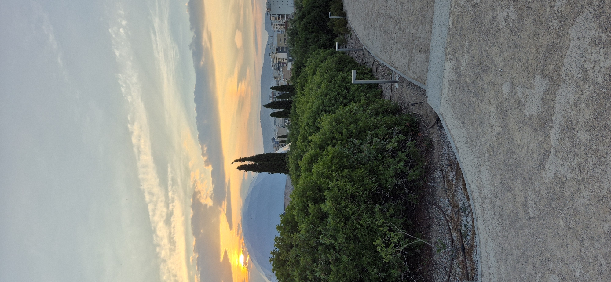 The image depicts an outdoor scene at dusk, likely in Kallithea, Greece, with a view of a large, white, dome-shaped structure partially obscured by lush green trees in the foreground. The sky is painted with hues of orange, yellow, and blue from the setting sun, with wispy clouds stretching across the horizon. In the mid-ground, several tall, slender trees stand out against a backdrop of city buildings and distant hills. A paved pathway curves through the scene, lined with modern, L-shaped lampposts, suggesting a park or public area. The ground is covered in gravel and some dry foliage. The overall impression is one of a peaceful, urban landscape at twilight.