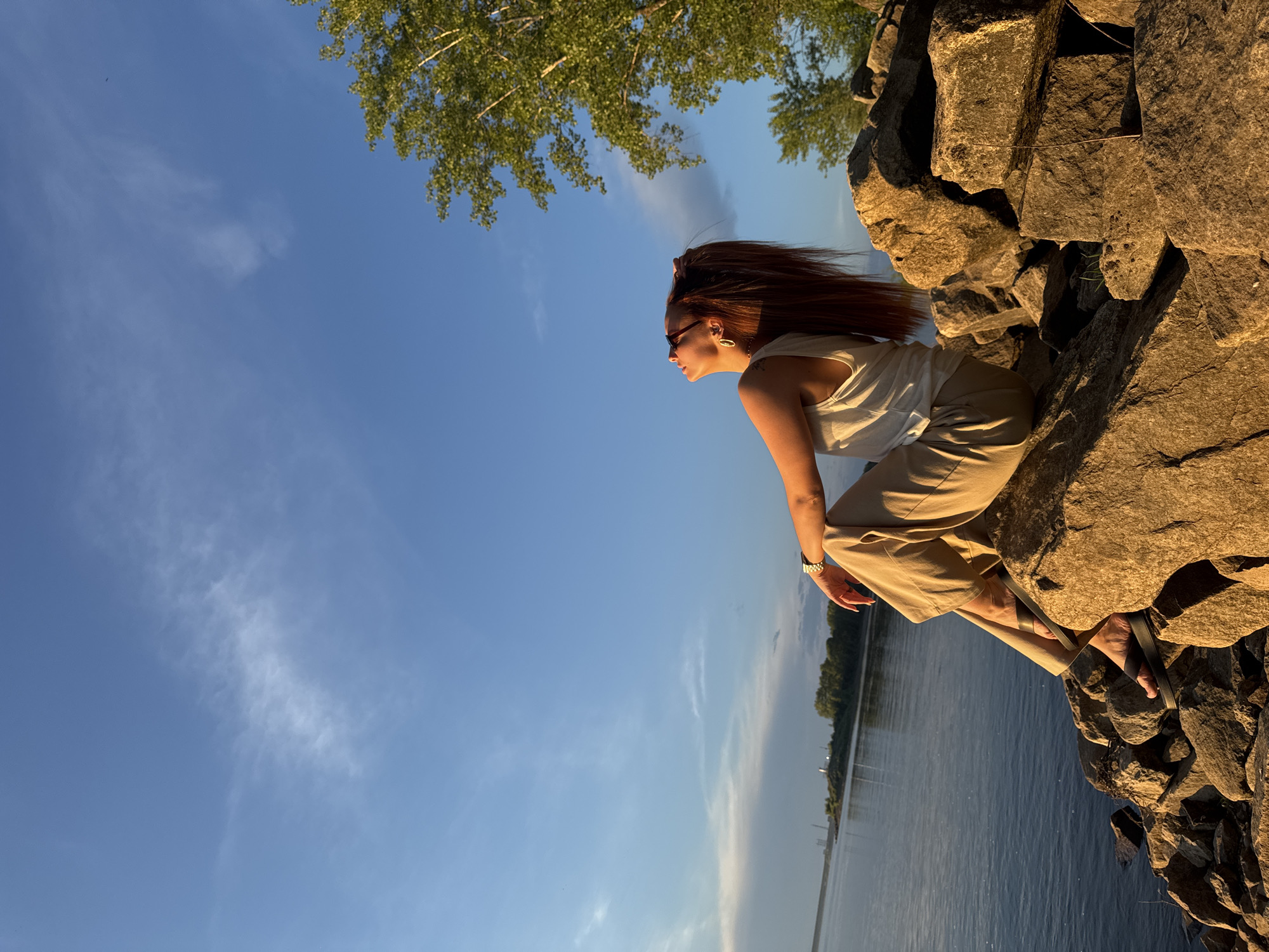 A young woman with long, auburn hair sits on a rocky shoreline, looking up at a clear blue sky with scattered clouds. She is wearing a white tank top and light-colored pants. The sun is setting, casting a warm golden glow on the rocks and the water. In the distance, across the water, is a tree-lined shore with some industrial buildings. The scene appears to be a peaceful moment by a river or lake, possibly the Kama River in Chaikovsky, Russia, known for its scenic beauty and industrial presence.