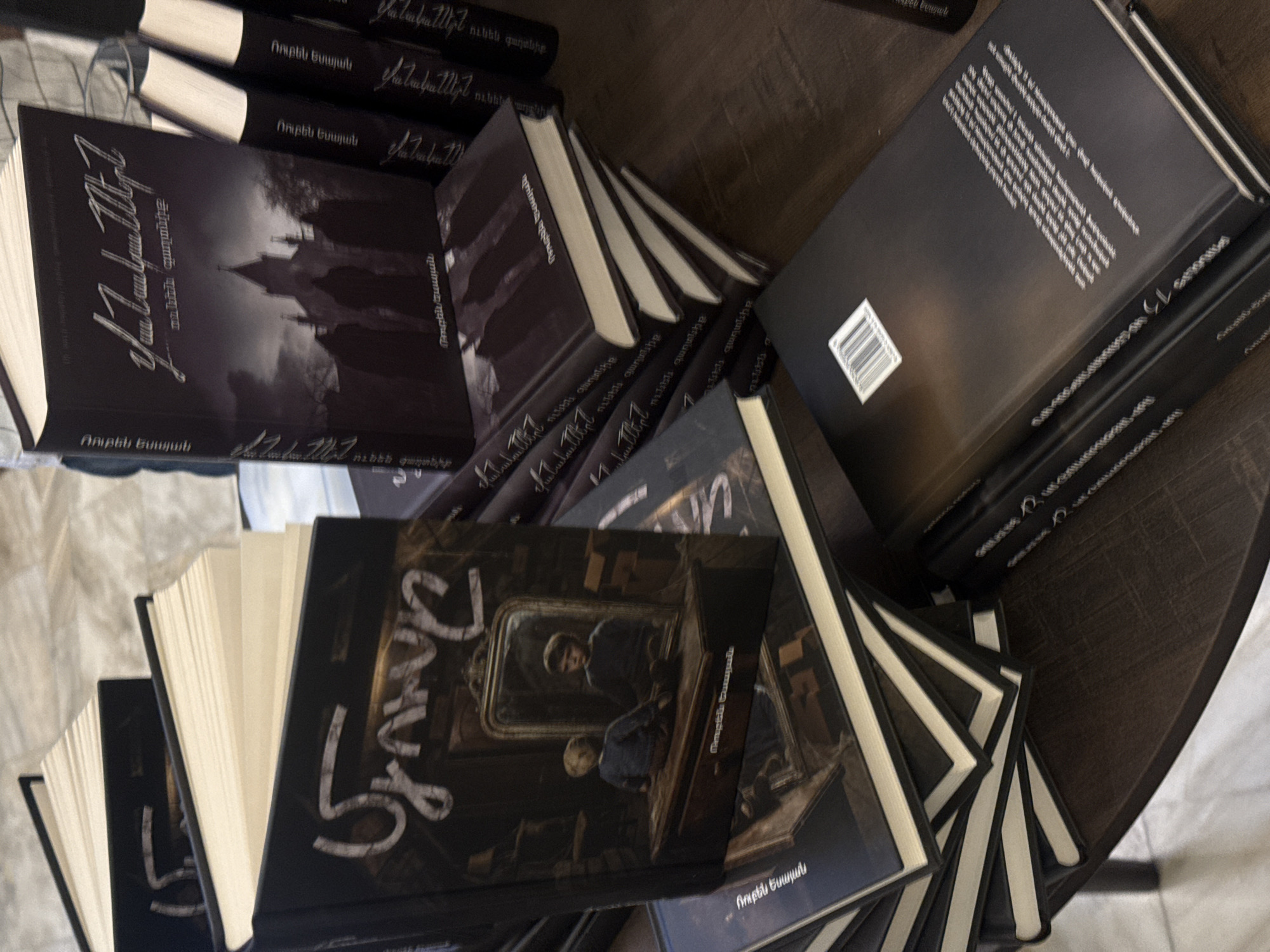 The image shows a collection of books stacked on a dark surface, likely a table. The books appear to be new and feature a dark, monochrome aesthetic. Two distinct book covers are visible: one with a gothic castle-like silhouette and the other with an image of figures reflected in a mirror inside a train car. The text on the covers is in Armenian, with the name "Ruben Yesayan" clearly visible on each. The location is likely a bookstore or library in Yerevan, Armenia, given the Armenian text.