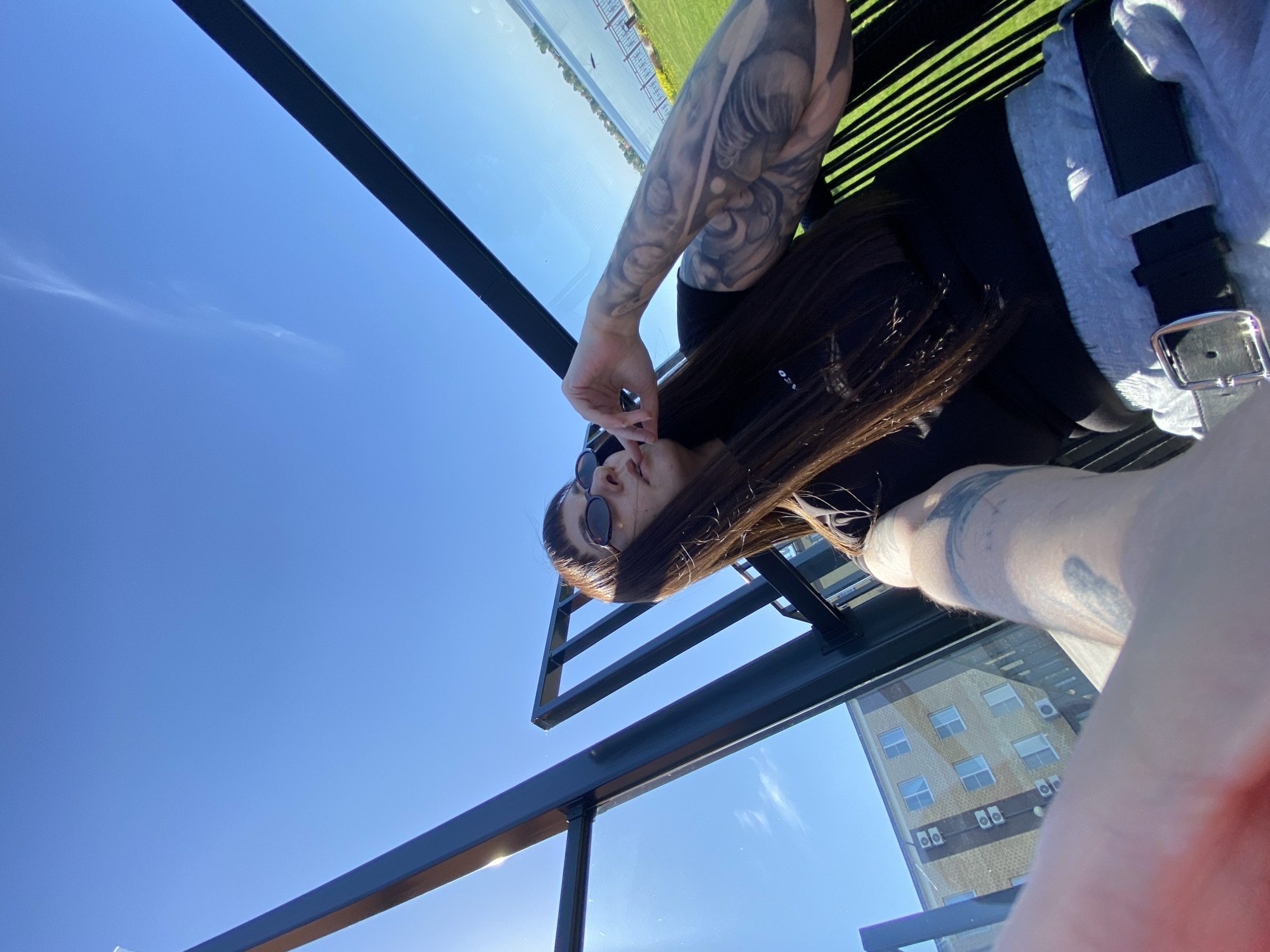 A selfie photograph of a person with brown hair wearing sunglasses, a black shirt, a grey shirt, and a black belt with a silver buckle is taken on a rooftop. The person has tattoos on their arms. The background features a bright blue sky, a distant grassy field, and a tall brick building with multiple windows and air conditioning units. The photograph appears to be taken in an urban area. The location in Votkinsk, Russia cannot be determined from the image.
