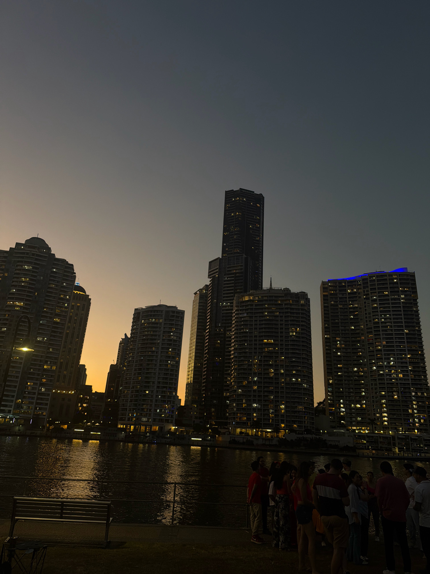 At dusk in Kangaroo Point, Australia, a group of approximately fifteen people are gathered on a paved walkway along a river. They are facing towards the water, and some appear to be engaged in conversation. Across the river, a cityscape is illuminated by the warm glow of twilight and the lights of numerous high-rise buildings. The water reflects the building lights, creating a shimmering effect. In the foreground, a metal park bench sits empty on a grassy area. The sky is a gradient of dark grey to pale orange, indicating the transition from day to night.