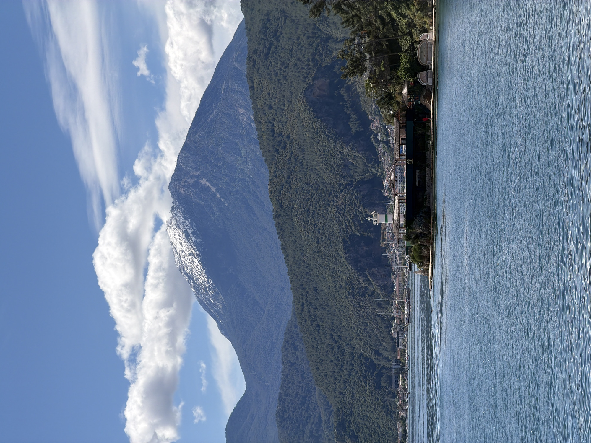 The image showcases a mountain range alongside a body of water, likely the Mediterranean Sea, in Fethiye, Turkey. The mountains are covered in dense green forests, transitioning to a rugged, rocky terrain towards the peaks. A line of buildings and boats lines the shore between the mountain and the water. The sky is partly cloudy, with scattered white clouds against a blue background.