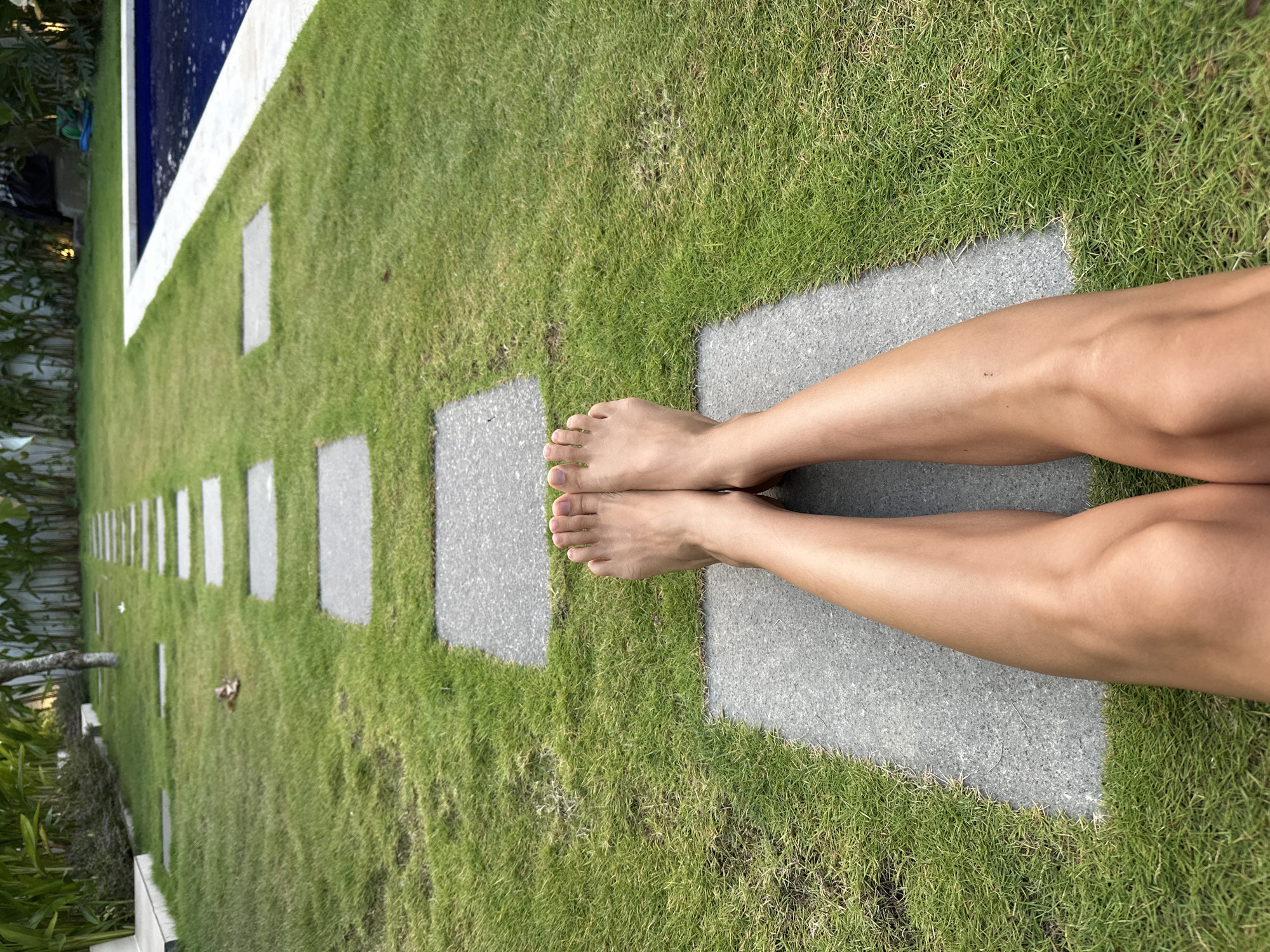 A low-angle shot shows a person's legs and feet resting on square stone pavers in a grassy area, with a swimming pool visible in the background. The scene appears to be in a resort or private residence in Kuta Selatan, Indonesia, suggested by the lush tropical vegetation and the style of the poolside area. The grass is a vibrant green, and the stone pavers are evenly spaced, creating a path. The swimming pool is a deep blue, and its edge is lined with white stone. The lighting suggests it is daytime, possibly late afternoon, with the sun casting a warm glow on the scene.