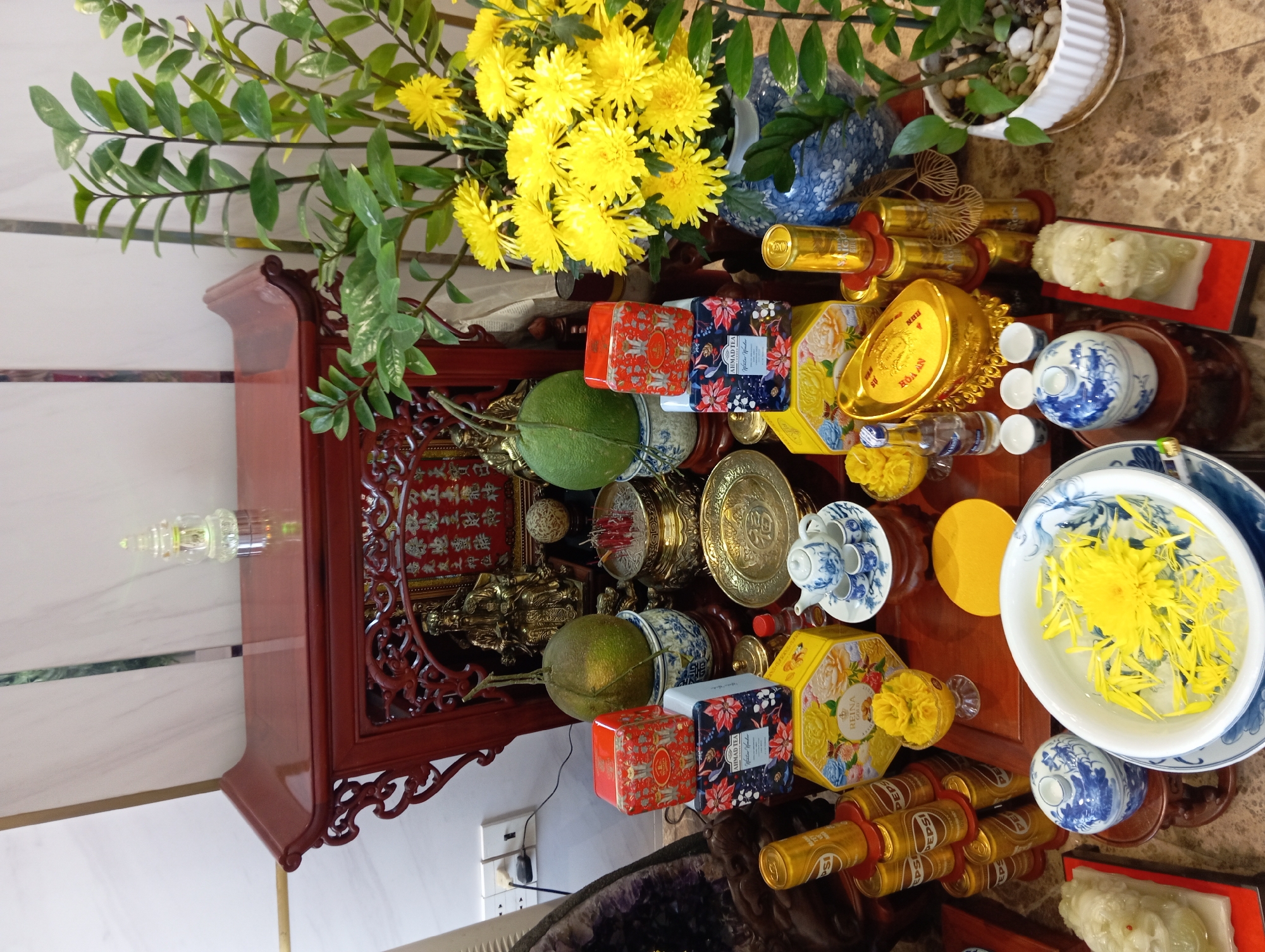 The image shows a wooden altar with various items arranged on it. The altar is adorned with intricate carvings and has a dark red finish. On the altar, there are two green fruits, likely pomelos, placed on decorative blue and white ceramic vases. Several stacks of cylindrical cans, possibly beverages, are visible, some bearing the "Pepsi" logo. There are also boxes of tea, one with a yellow floral design and the brand "Reina Gold" visible. A small blue and white teapot with matching cups sits on a plate. Other decorative items include golden objects shaped like ingots, a small bottle, and what appear to be white figurines. A cluster of vibrant yellow chrysanthemums in a blue and white vase adds a splash of color to the scene. The altar is placed on a marble-patterned surface. In the background, a plant with green leaves grows upwards, and a portion of a white wall with electrical outlets is visible. The overall impression is that of an offering or a shrine, possibly for a special occasion or cultural ritual. The presence of the "Saigon Gold" cans suggests a Vietnamese context, and the overall arrangement is consistent with household altars often found in Vietnam, including in cities like Nha Trang.