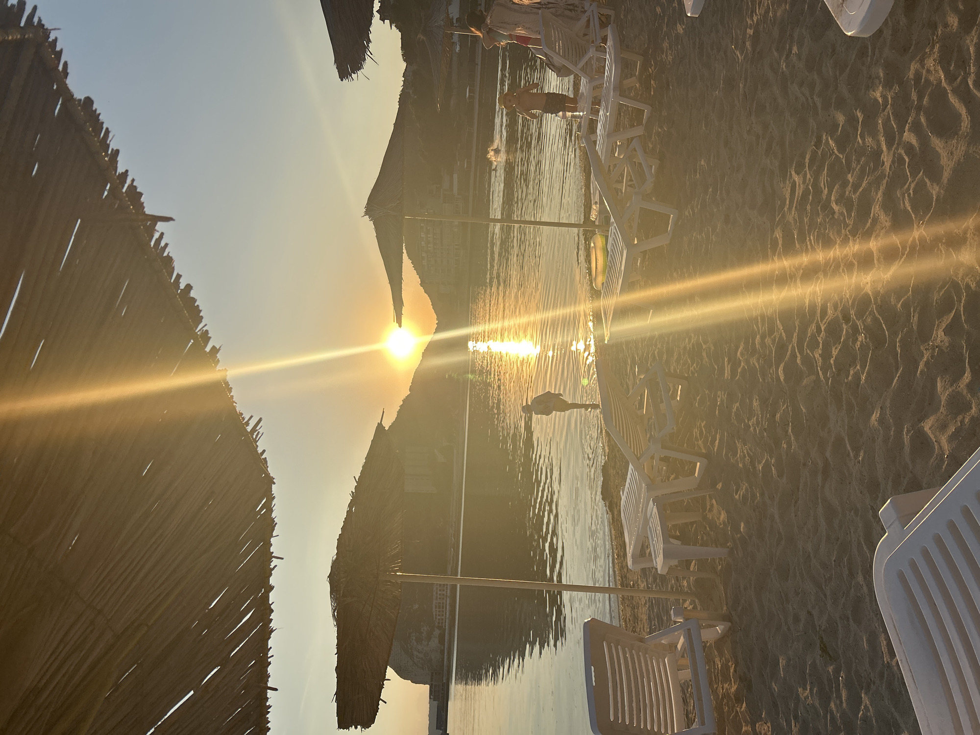 This is a photograph of a beach in Kavarna, Bulgaria, during sunset. The sun is low on the horizon, casting a warm, golden light across the scene. The beach is made of sand, and there are several white lounge chairs and a small table set up, suggesting a tourist resort. In the foreground, the straw roofs of two beach umbrellas are visible, with one umbrella prominently featured. The sea is calm, with gentle waves lapping at the shore. In the distance, a rocky cliff face with buildings is visible, and the sun's reflection shimmers on the water. There are a few people in the water, including what appears to be two children. One child is standing near the shore, and another is further out in the water. A woman is standing near some lounge chairs, and another person is partially visible behind her. The overall atmosphere is peaceful and serene, capturing the beauty of a sunset at the seaside.