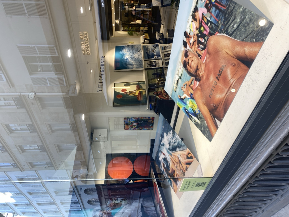 This image depicts the display window of a YellowKorner photography gallery located at 10 Rue de Solférino, 75007 Paris, France.
The scene is viewed from an outdoor street perspective looking into the gallery window. In the foreground, three large, glossy photographic prints are prominently displayed. The print on the right shows an elderly, tanned male with white hair and sunglasses, reclining on a beach chair, with the phrase "TUTTO PASSA" tattooed on his chest. Other individuals and colorful beach chairs are visible in the background on a rocky shoreline next to calm blue water. The central print features a close-up of multiple sets of legs and feet positioned above turbulent blue ocean water, suggesting a boat or pier setting. The leftmost print is partially obscured, showing a black and white image, possibly a portrait or a structured scene. A green informational sign with a QR code and the text "Rendez-vous photographique!" is positioned on the display surface near the left print. A dark gray metal railing, part of the building's facade, is visible along the bottom edge of the window.
Through and reflected in the glass, the interior of the YellowKorner gallery is visible, featuring additional framed photographs on walls and pedestals under bright interior lighting. A prominent "YELLOW KORNER" sign is affixed to the back wall. Other visible artworks inside include a picture of a woman on a yellow inflatable object holding red balloons, and a dynamic blue and white image resembling ocean waves. A reversed reflection of a sign stating "THE FRENCH BASTARDS" is also apparent.
The reflections on the glass also reveal the exterior environment across the street. These include typical Parisian Haussmannian buildings with light-colored stone facades, multiple stories, regularly spaced windows, and white louvered shutters. A reflected street sign, "RUE DE SOLFÉRINO," is discernible in reverse on the upper right side, confirming the location. Faint reflections of people and other street elements are visible on the left side of the window. The overall lighting suggests a bright, possibly sunny, daytime condition.