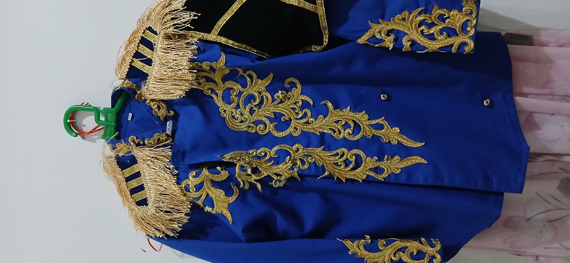 The image shows a blue ceremonial jacket with gold trim and fringe, hanging on a green plastic hanger against a white wall. The jacket has ornate gold embroidery on the front and sleeves, and gold fringe along the shoulders. A black fabric panel with gold trim is visible on the right sleeve. A pink floral-patterned fabric is visible underneath the jacket. The jacket appears to be a costume or uniform. The scene is indoors, with the wall being the only discernible environmental feature. There are no people or animals in the image. There is no information in the image to indicate the city of Samarinda, Indonesia.