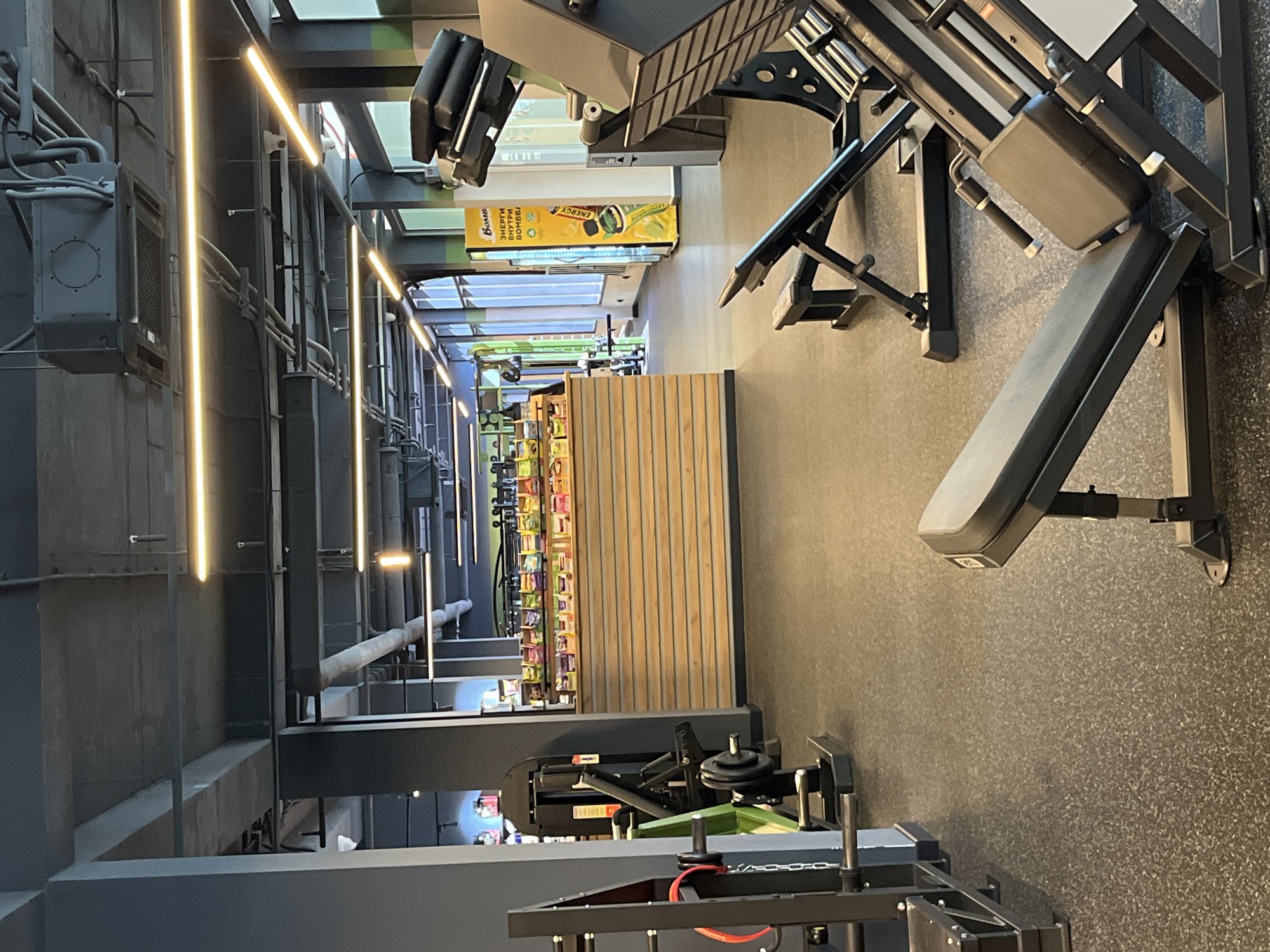 This image depicts a modern, unoccupied indoor gym facility located in Sochi, Russia. The scene is characterized by a dark industrial aesthetic, featuring an exposed ceiling painted dark grey with visible pipes, air ducts, and concrete beams. Linear LED strip lights provide illumination, casting a bright, warm glow throughout the space.
The flooring is a black speckled rubber material typical of gyms. On the right side of the foreground, several pieces of black metal gym equipment are present, including an incline leg press machine with black padded seats and a separate adjustable utility bench.
In the mid-ground, slightly to the left of the center, a wooden-slatted counter or kiosk is prominently displayed. Its top surface holds numerous colorful packaged snack items, likely protein bars or supplements. Behind this counter, additional products are visible on shelves through an opening. To the left of the counter, partially obscured, is another piece of gym equipment, possibly a weight rack, featuring a green accent and a black weight plate.
In the background on the right, a tall yellow vending machine is visible, displaying "BOMB" at the top and Cyrillic text that translates to "ENERGY INSIDE THE BOMB," consistent with a Russian brand. Large windows line the right wall, allowing natural light into the facility. No individuals are present, and no actions or interactions are depicted within the scene.
