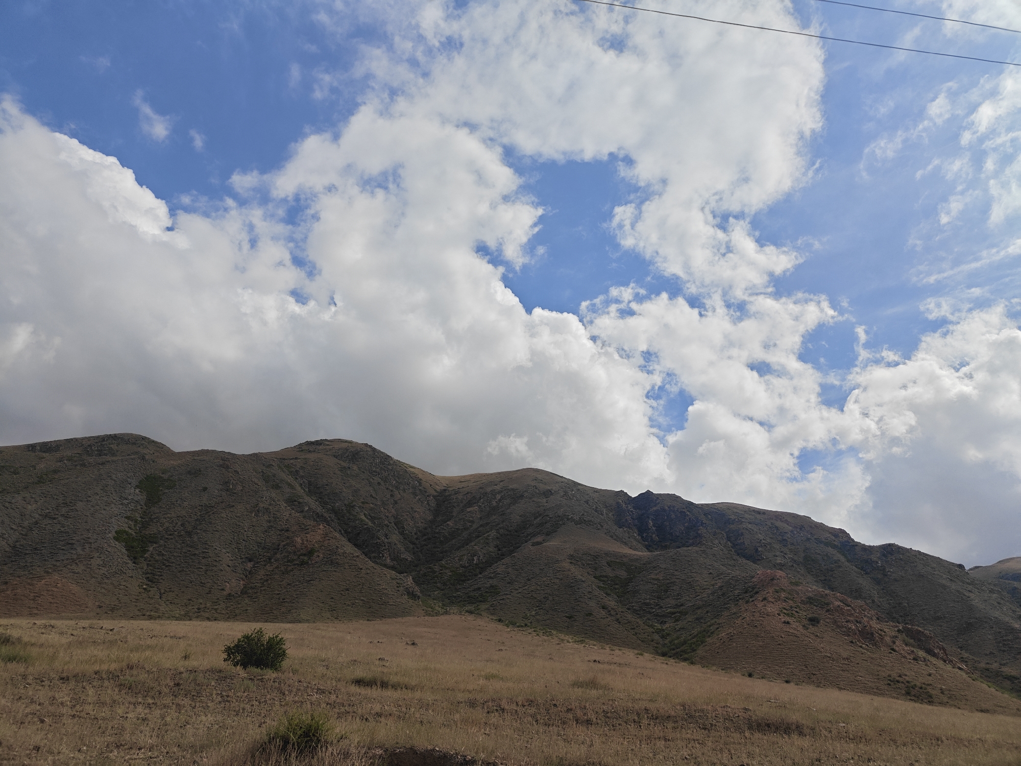 The image depicts a scenic view of rolling hills and mountains under a bright blue sky filled with fluffy white clouds. In the foreground, dry, golden grass covers the flat terrain, with a solitary green bush adding a touch of color. The hills are covered in sparse vegetation, with darker patches possibly indicating shadows or denser plant growth. The sky is a vibrant blue, with a network of power lines stretching across the upper portion of the frame. The overall impression is one of a vast, open natural landscape. The location is identified as near Kadji Say, Kyrgyzstan.