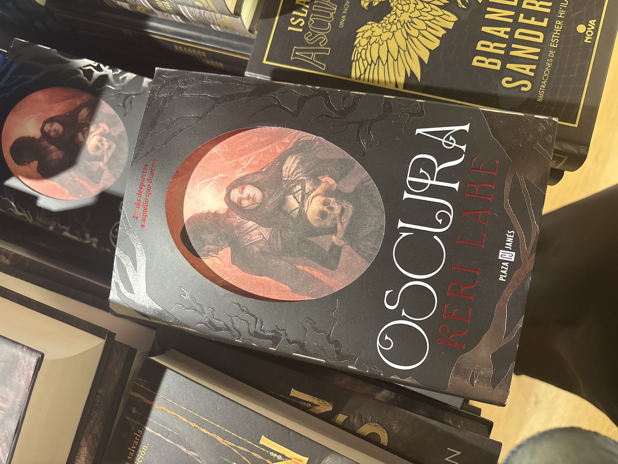 A close-up, angled shot shows several books on display, with a focus on the book titled "OSCURA" by KERI LAKE. This book has a black cover with a circular cutout revealing an image of a woman holding a skull, silhouetted against a red background. The title is in white, stylized lettering, and the author's name is in red. To the right of "OSCURA," another book is visible with a black cover and gold lettering that reads "ISLA ASCUA UNA NOVA." Further to the right, a portion of another book is visible with gold lettering that spells out "BRANDON SANDERS." The background appears to be a bookstore or library setting. The lighting is somewhat dim, with a spotlight effect on the books. The image does not provide information about the city of Orihuela, Spain, or any specific locations within it.