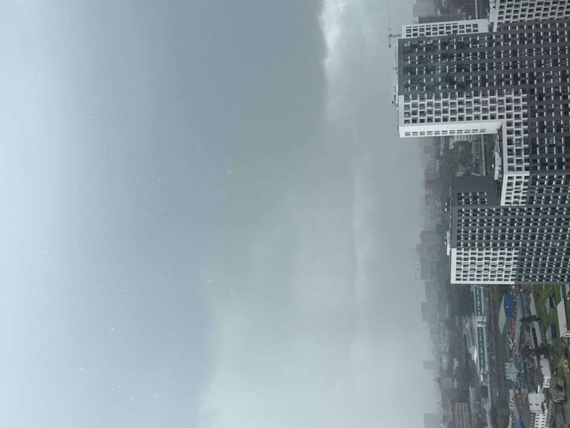 The image depicts an overcast sky with a hint of precipitation in the form of tiny white specks. In the foreground, two modern high-rise apartment buildings dominate the scene, characterized by their alternating dark and white facades with numerous rectangular windows. The building on the right is taller and more slender than the one on the left, which is broader and appears to be composed of multiple connected sections.
Beyond these prominent structures, the middle ground reveals a sprawling cityscape of Lyubertsy, Russia. This area consists of a dense collection of various buildings, including residential blocks, industrial facilities with blue and orange containers, and what appear to be construction sites with cranes visible in the distance. The overall atmosphere is hazy, likely due to the weather conditions, with the background city fading into the atmospheric haze. Trees and greenery are interspersed throughout the urban landscape, suggesting a mix of residential and industrial zones within the city. The sky is a uniform dull gray, contributing to a subdued and potentially somber mood. There are no visible people in the image.
