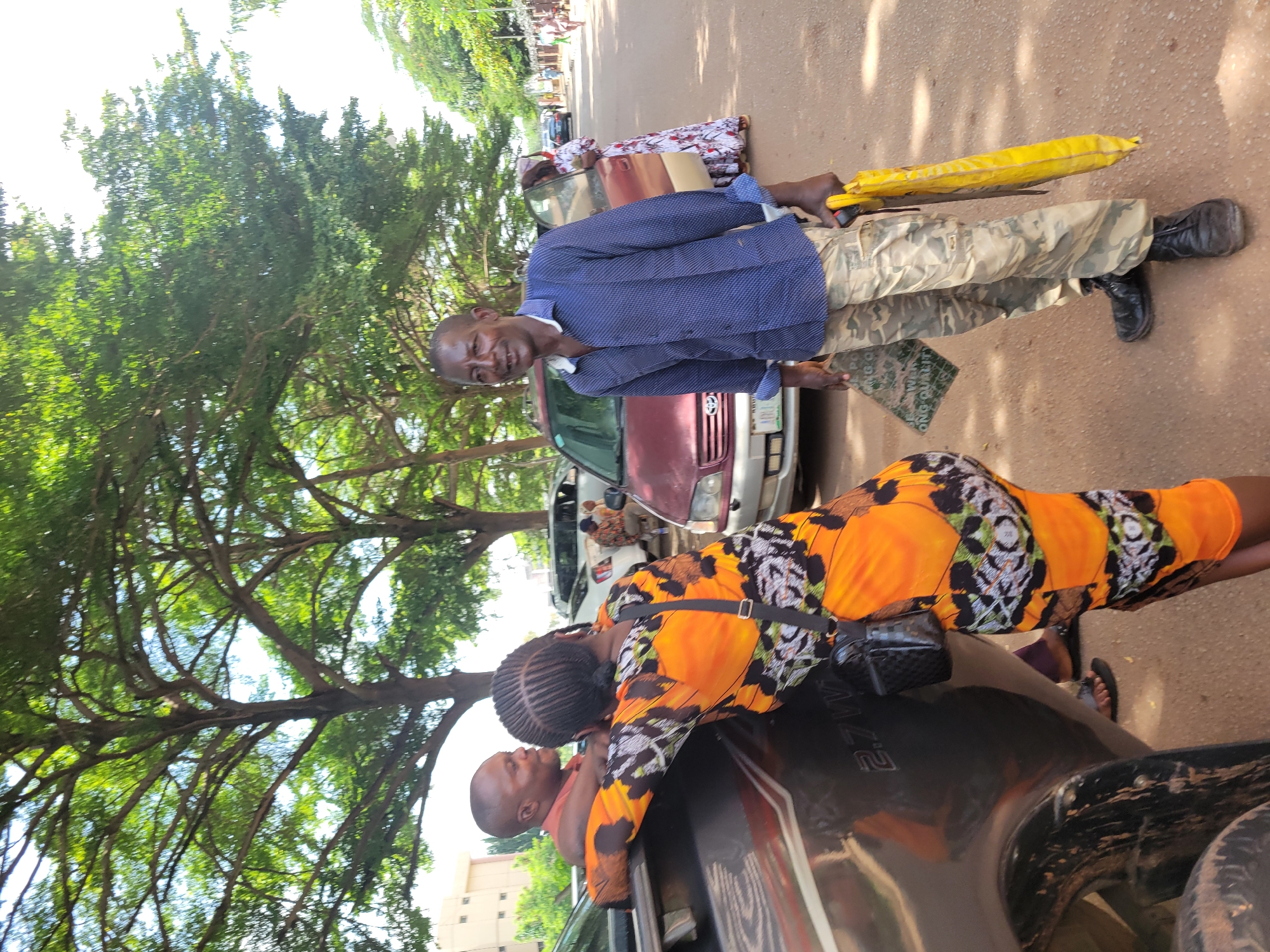 This outdoor image, taken in Abuja, Nigeria, captures a vibrant street scene under bright daylight. The setting is a paved road lined with lush green trees, indicating a verdant urban environment.
In the foreground, a man stands smiling slightly at the camera. He is dressed in a blue long-sleeved, collared shirt, camouflage-patterned trousers, and dark boots. He holds a bright yellow, possibly folded umbrella or package, in his right hand. Near his left foot, a flat green object with partially visible text, reading "ON GO QUALI," rests on the ground.
To the man's right, a woman with braided hair, wearing a vivid orange, black, and white patterned dress, leans into a dark-colored vehicle, appearing to converse with a person inside. A small black fanny pack is visible around her waist. The head and upper torso of a man in a red shirt can be seen inside the vehicle, interacting with her. Text, possibly "2.7V" or "2.7L," is faintly visible on the side of this vehicle.
In the mid-ground, a silver Toyota SUV is parked behind the main man, and further back, a reddish-brown car with an open door is visible, near which another person in a long white patterned dress stands. The bright, clear weather is evident from the strong overhead light and shadows. The scene suggests a casual, everyday interaction or a brief stop along a busy street, with the subjects appearing relaxed.