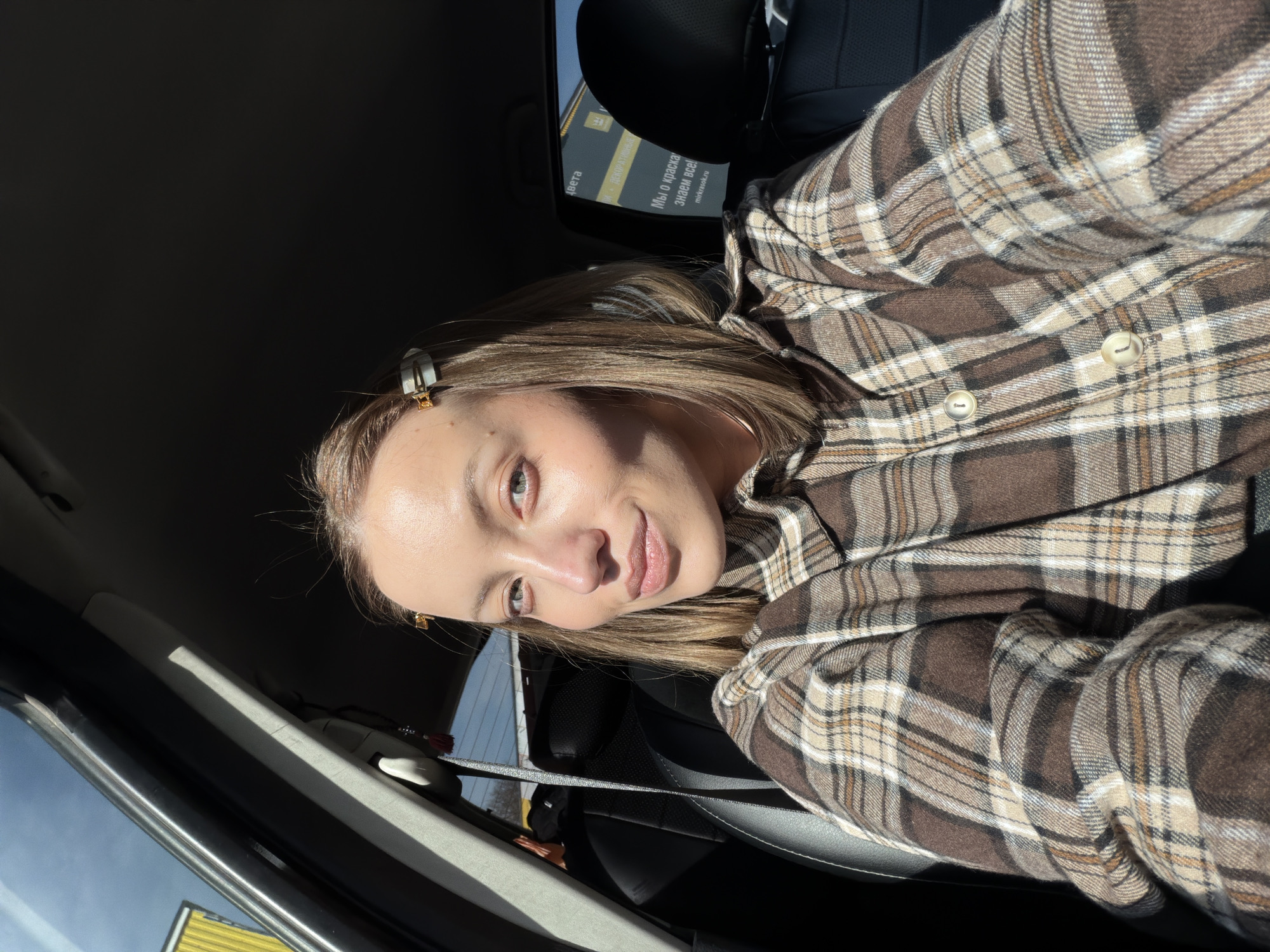 A young woman is taking a selfie from inside a car. She is wearing a brown and beige plaid shirt. She has blonde hair pulled back with clips. The car's interior is dark, and sunlight is shining in through the window. A sign is visible through the car window, and the text "ШИ О КРАСКА ЗНАЕМ ВСЁ" and the website "mirkrask.ru" are readable, indicating a business possibly related to paint. The woman appears to be in Shevlyakovo, Russia, based on the website suffix.