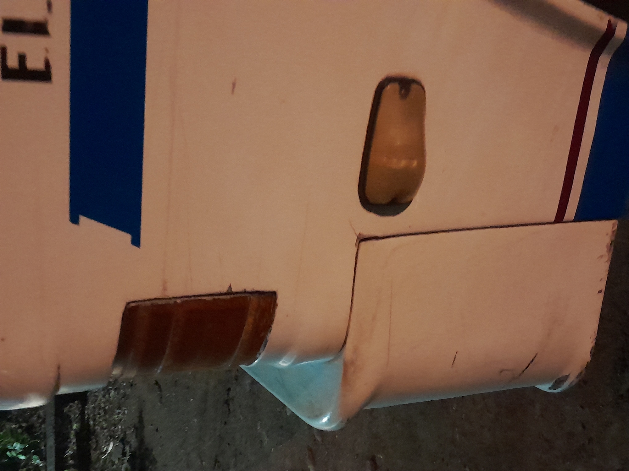 The image shows a close-up of the rear quarter panel of a white vehicle, possibly a bus or a large truck, parked on a dirt or gravel surface. The vehicle has a blue stripe running horizontally across its side, with a thin red stripe above it. A reflector or indicator light is visible on the rear quarter panel, and a brake light assembly is present above it. Part of the word "EL" is visible in black lettering at the top right of the vehicle. The background is blurred, showing dark foliage and the ground. The scene appears to be in an outdoor, dimly lit environment, possibly at dusk or night. The presence of this specific vehicle type and the visible lettering suggests a location in Santa Ana, Venezuela, as it's a common vehicle for public transport in that region.