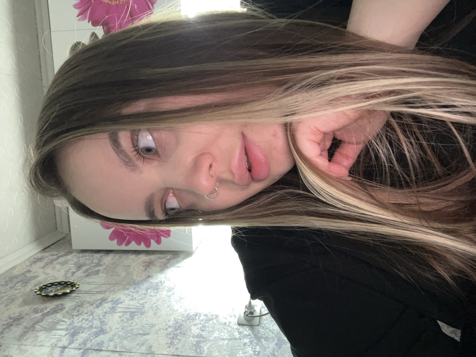 A woman with brown and blonde highlighted hair rests her face on her hand. She has a silver hoop nose piercing. She is wearing a black top. The background shows a patterned floor, a white cabinet with a pink flower design, and a wall. An electrical outlet and a decorative object are visible on the floor. The location is potentially Novopokrovskaya, Russia, although this is not definitively confirmed by the image.