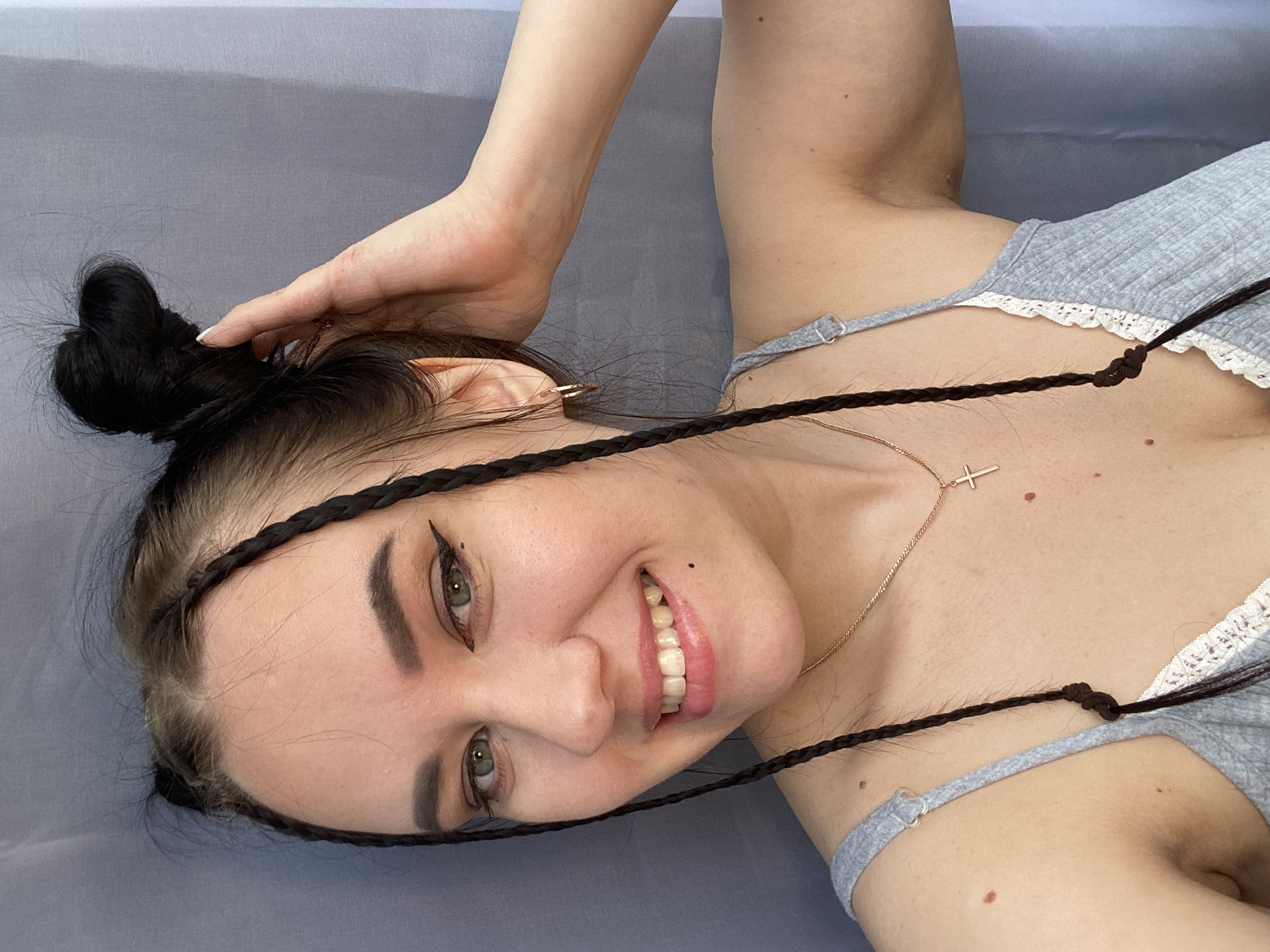 A young woman with fair skin, dark hair styled in a bun and two braids framing her face, is lying on a gray surface. She is wearing a light gray tank top with white lace trim and a delicate gold necklace with a cross pendant. She smiles, revealing her teeth. The location and city of Chaikovsky, Russia, are not evident from the image.