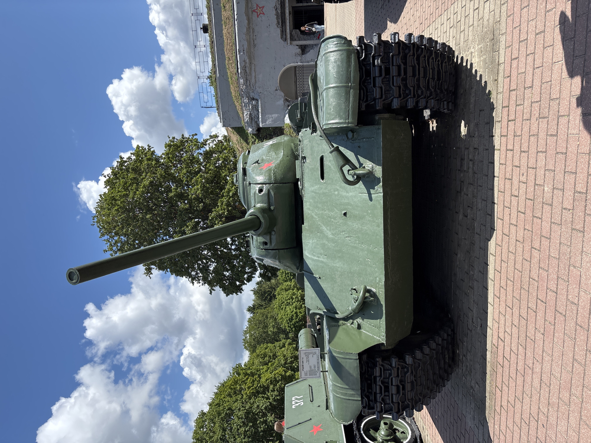 In Brest, Belarus, a prominently displayed Soviet IS-2 heavy tank is positioned on a red brick paved area, likely within a museum or memorial complex. The tank is painted in a matte green military camouflage. Its large gun barrel points directly forward, elevated slightly. Red stars, a common Soviet military insignia, are visible on the turret and on another smaller tank or vehicle situated to the left of the IS-2. This auxiliary vehicle also bears the number "327" and a label that reads "СРЕДНИЙ ТАНК Т-34" which translates to "Medium Tank T-34", indicating its classification.
Behind the tanks, a fortified structure with a grassy, sloped roof rises. This structure appears to be part of a historical fortification, possibly a fort, given the presence of embrasures and an arched entrance. A dark metal railing runs along the top of the fortification.
The sky above is a bright blue, dotted with fluffy white clouds. Lush green trees form a dense backdrop to the scene, suggesting a summer day. A few people are visible in the distance near the entrance of the fortified structure, one of whom appears to be a woman standing and looking into the building. The overall impression is one of a static display of military hardware against a historical and natural background.