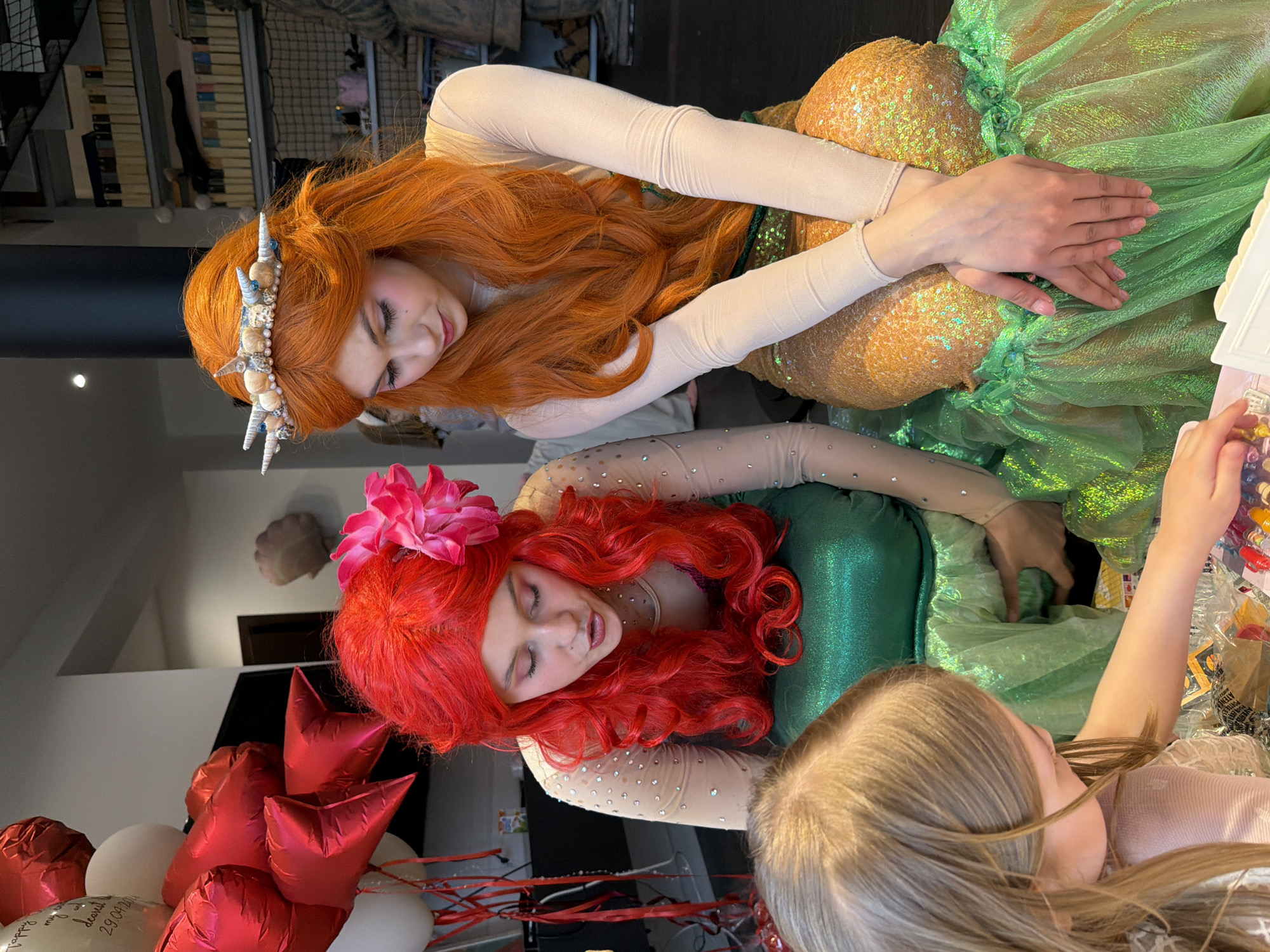 In a bright indoor setting, two individuals costumed as mermaids are positioned vertically, attending to a child visible in the lower right corner. The upper mermaid has long, red-orange hair adorned with a seashell crown, and wears a shiny gold and green mermaid tail. The lower mermaid has bright red, curly hair with a large pink flower, and a shiny green mermaid tail. Both are wearing light beige, long-sleeved tops with small embedded gems. To the left are several red heart-shaped and white round balloons. The location is Крючково-2 Снт, Russia.