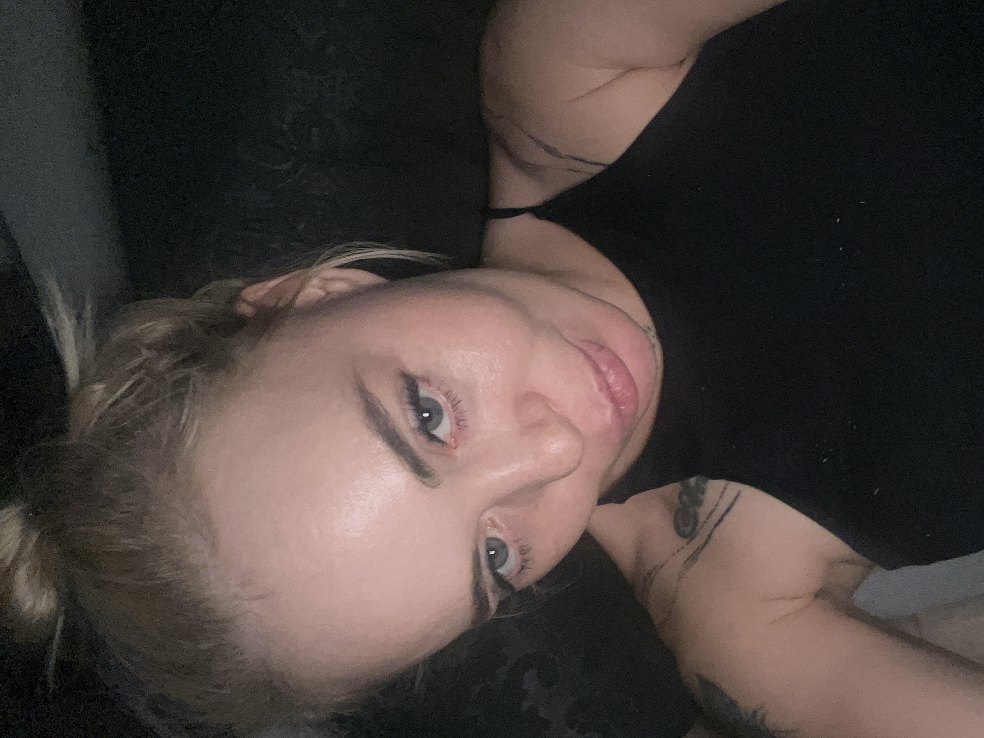 In a dimly lit, indoor environment, a woman with blonde hair pulled into a bun is captured in a close-up, frontal selfie. She is wearing a black halter top, and her bare shoulders and upper chest are visible, adorned with tattoos. The woman's face is tilted slightly downwards, and she appears to be looking towards the camera with her blue eyes. Her lips are slightly parted, and she has makeup on, including mascara and possibly eyeliner. The background is dark and indistinct, with some subtle texture suggesting a wall or fabric. The lighting in the image is low, casting shadows and highlighting certain features of her face.
