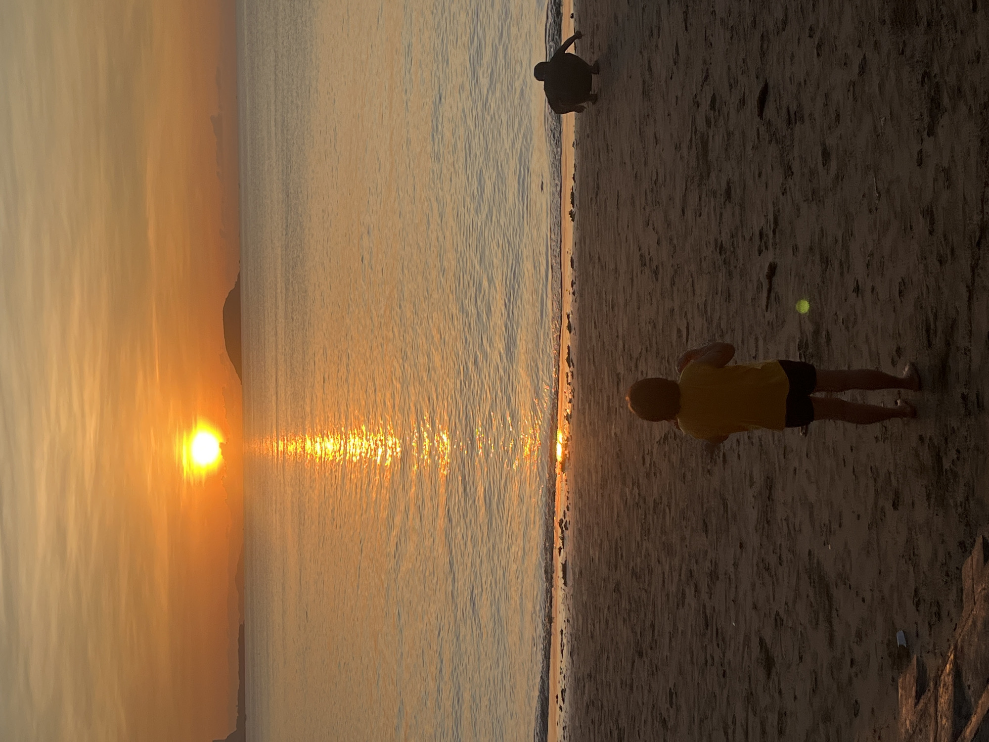 The image depicts a beach scene at sunset in Bac Nha Trang Ward, Vietnam. The sun is setting over the ocean, casting a golden light. The water reflects the light, creating a bright reflection. In the foreground, a child in a yellow shirt and dark shorts stands on the sandy beach. Further up the beach, another person is crouched. The beach is dark and sandy. There is a wall at the end of the beach.