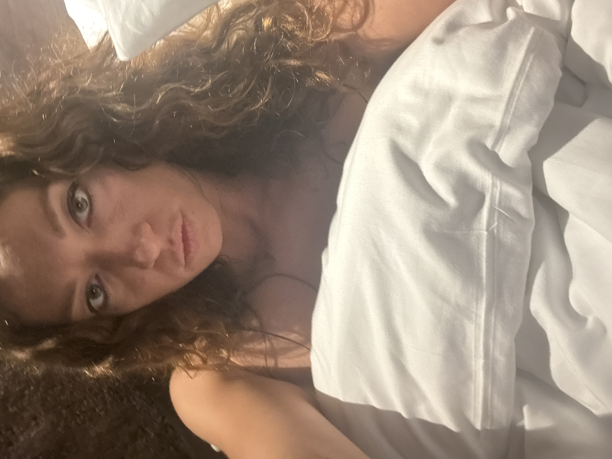 A close-up, overhead shot shows a woman with wavy brown hair lying in bed. She is partially covered by a white duvet. The woman's eyes are open and she is looking directly at the camera. Her expression is neutral. The background is dark and out of focus.