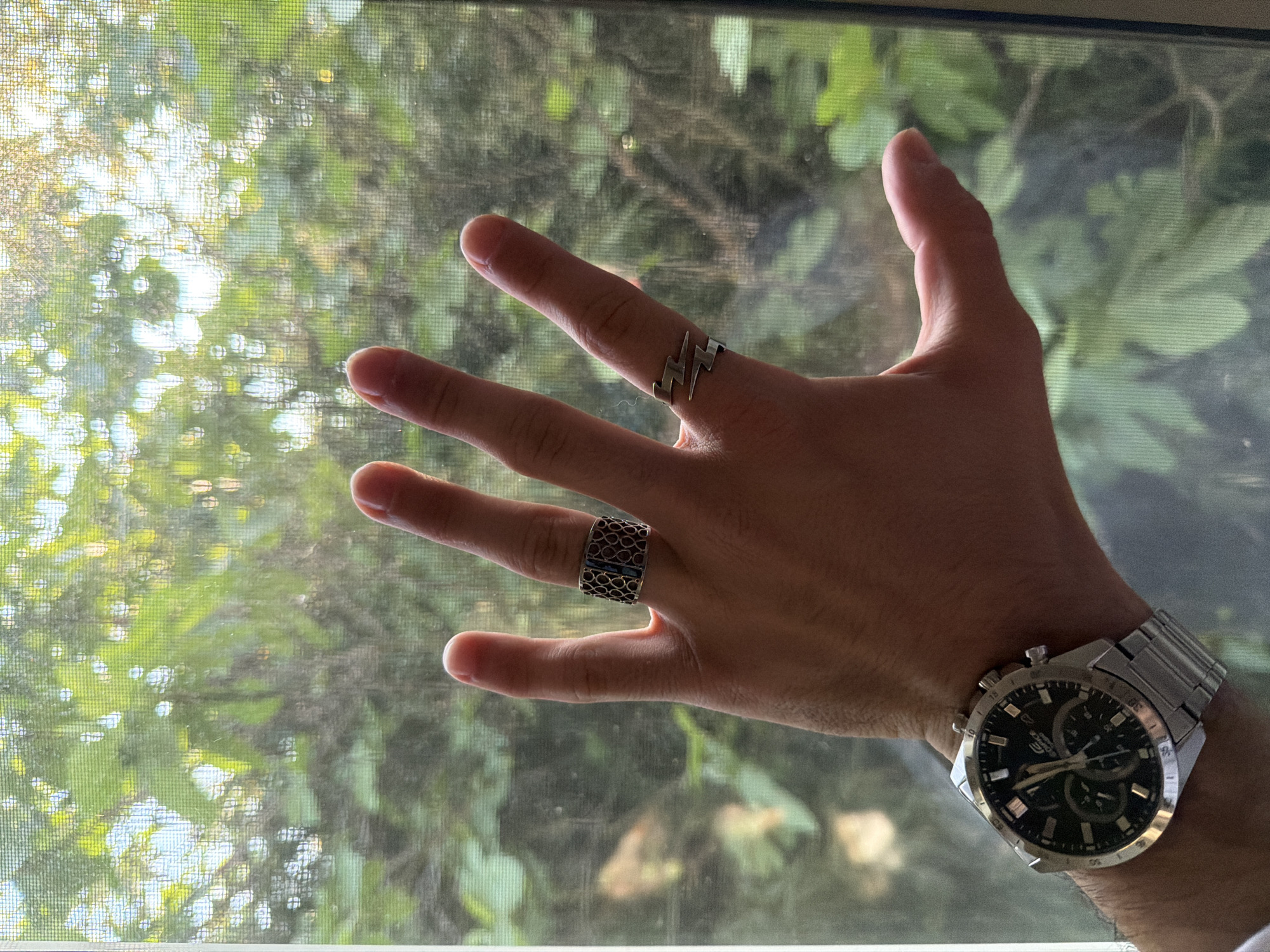 A hand with a silver watch and two rings is held up in front of a window. The ring on the ring finger is a wide band with a geometric pattern, and the ring on the middle finger is a silver band with a lightning bolt design. The watch is silver with a black face. The window is covered with a screen, and the background is blurred greenery. It is impossible to know the location of the image based on the given content.