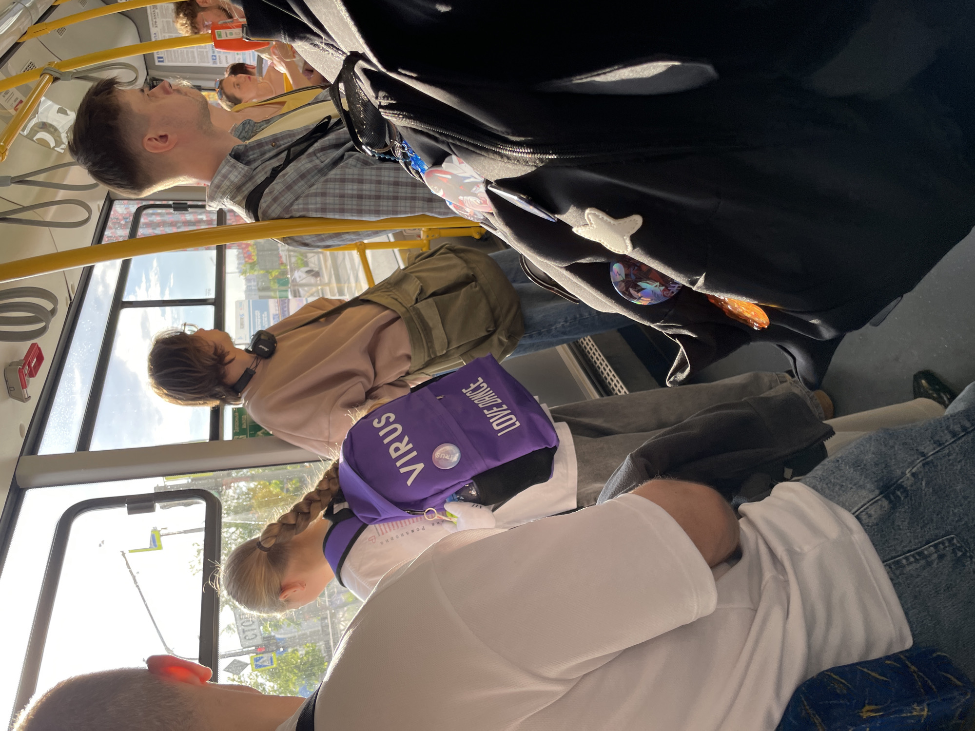 The image depicts the interior of a public transport vehicle, likely a bus or trolleybus, in Saint Petersburg, Russia, during daytime under clear, sunny weather. The scene is captured from a low, upward-looking perspective and is rotated clockwise.
Multiple passengers are visible. In the foreground, the back of a person's head and shoulder, wearing a white shirt, is seen. Adjacent is a young person with blonde or light brown hair in a braid, also in a white top, carrying a bright purple backpack. The backpack distinctly features the large white text "VIRUS" and "LOVE DANCE," along with a circular pin also reading "VIRUS."
Further back, a person wearing a light brown or pink top has headphones around their neck and carries an olive-green backpack. Behind them, a man in a plaid shirt is looking upwards and forward. A large black backpack, adorned with several decorative pins and badges, including one shaped like a white star and others with iridescent designs, is partially visible, likely belonging to a passenger.
The vehicle's interior features characteristic yellow grab bars and hand loops. Through the windows, strong sunlight illuminates the scene, casting highlights and shadows. The urban environment outside is partially visible, showing glimpses of street signs with Cyrillic text and distant buildings, consistent with Saint Petersburg. Passengers appear to be passively commuting, and no specific activities beyond riding are apparent. One partially visible Cyrillic sign inside the vehicle is red and likely informational.