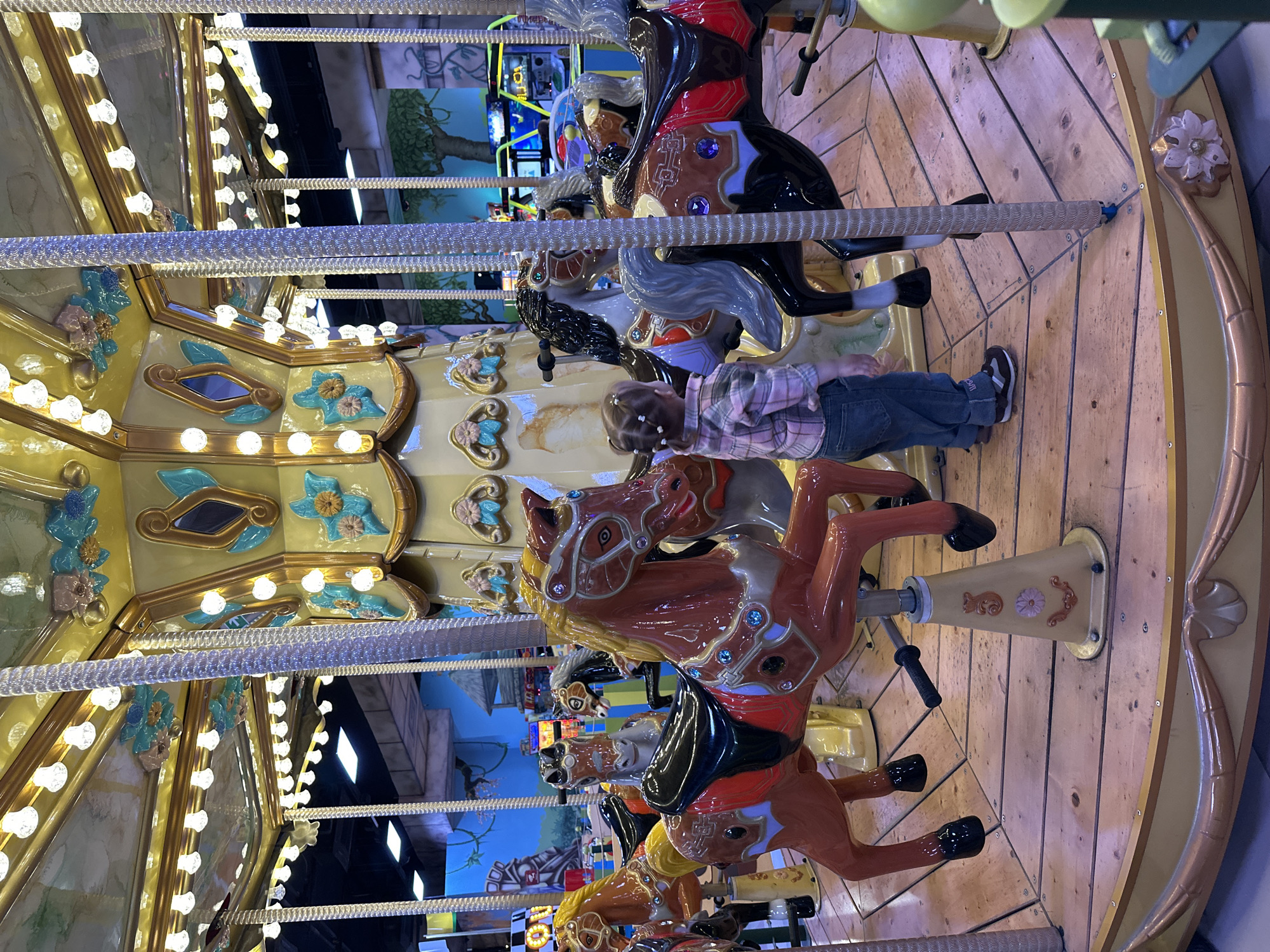 The image shows a carousel with multiple painted horses. A child is riding one of the horses, wearing a pink plaid shirt, blue jeans, and sneakers. The carousel is decorated with lights and ornate carvings. The background shows a mural depicting trees and other fantasy scenery. The location is inferred to be in the city of Yekaterinburg, Russia based on the objects.