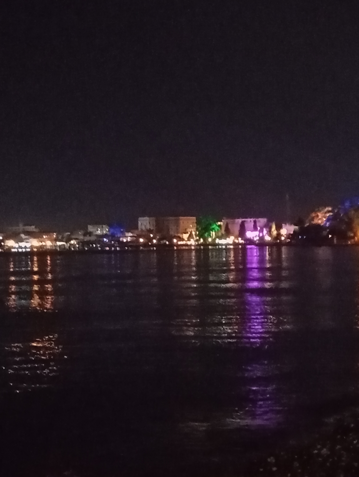 A nighttime, wide-angle, low-resolution photograph depicts a waterfront cityscape with illuminated buildings reflecting in dark water. The city appears to be Sokhumi, Abkhazia, with a prominent, multi-story building structure in the mid-ground, lit with purple and green lights. Several other buildings and structures are visible along the waterfront, some with orange and white lights. The water in the foreground is dark and reflects the lights from the city, creating streaks of color. The sky is dark and clear. There are no people or discernible vehicles visible.