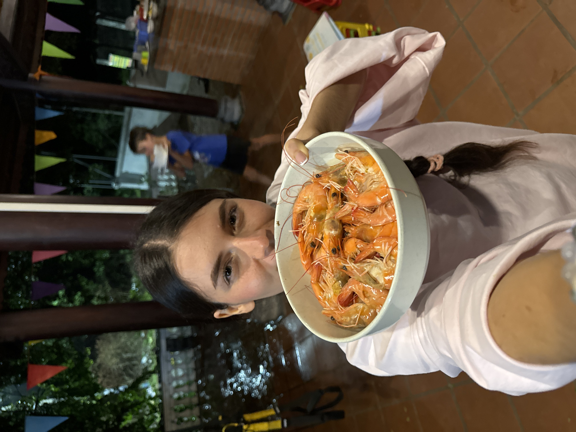 An eye-level portrait shot captures a young woman holding a large white bowl overflowing with cooked, reddish-orange shrimp. The woman, with dark hair tied back in a braid secured by a pink elastic, is wearing a light pink long-sleeved garment. She looks directly towards the camera with a neutral to slightly smiling expression. Her right arm and hand are visible, holding the bowl towards the viewer, with neatly manicured nails.
The setting appears to be outdoors at night or in the evening, indicated by the dark background and reflections on the floor. In the blurred background, another person, possibly a male, wearing a blue t-shirt and dark shorts, stands or walks further away, holding a white object. The ground is covered with terracotta-colored tiles, which show reflections of light sources, suggesting a wet or highly reflective surface. Overhead, colorful triangular bunting in green, yellow, and purple hues is strung up, hinting at a festive occasion or party. Dark foliage is visible behind the flags. The overall scene suggests a casual social gathering, likely in Mũi Né, Vietnam, where the shrimp are a central part of the meal or celebration.
