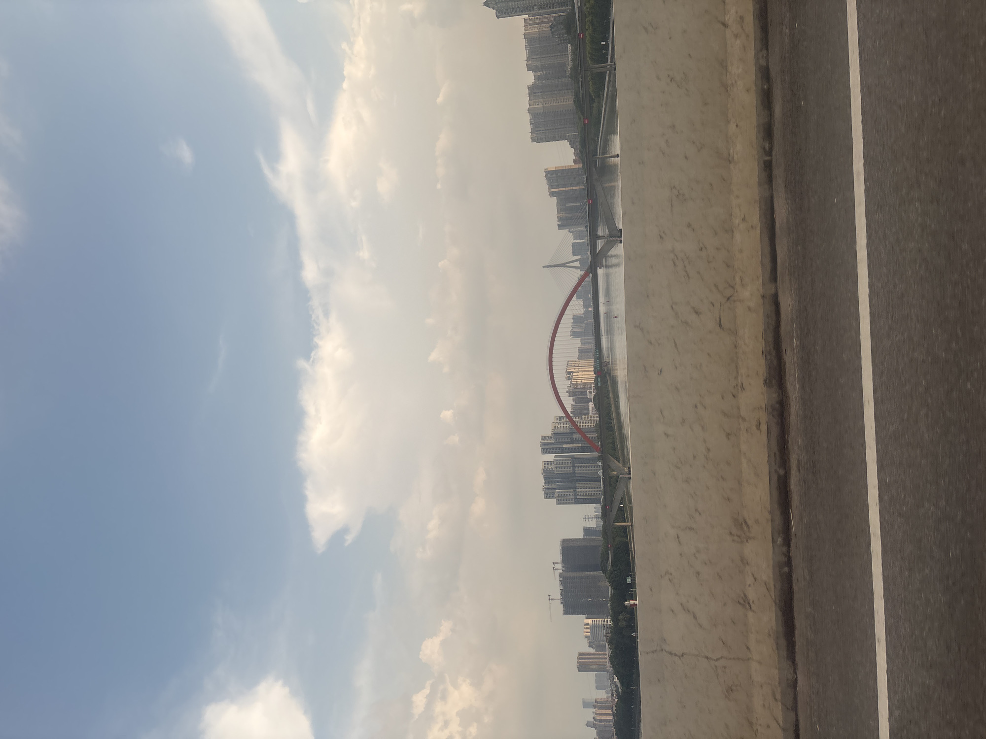 The image shows a cityscape with a river and a bridge in Baiyun District, China. The sky is partly cloudy with a mix of blue sky and white clouds. In the foreground, there is a concrete barrier and a road with a white line. Beyond the barrier, a wide river reflects the sky. On the opposite bank of the river, a dense urban area is visible, characterized by numerous tall residential buildings and some commercial structures. In the middle of the cityscape, a prominent red-arched bridge spans the river, with a cable-stayed bridge visible further in the distance. Several cranes are also present, indicating ongoing construction in the area. The overall scene depicts a modern city environment with its infrastructure and residential areas situated along a waterway.