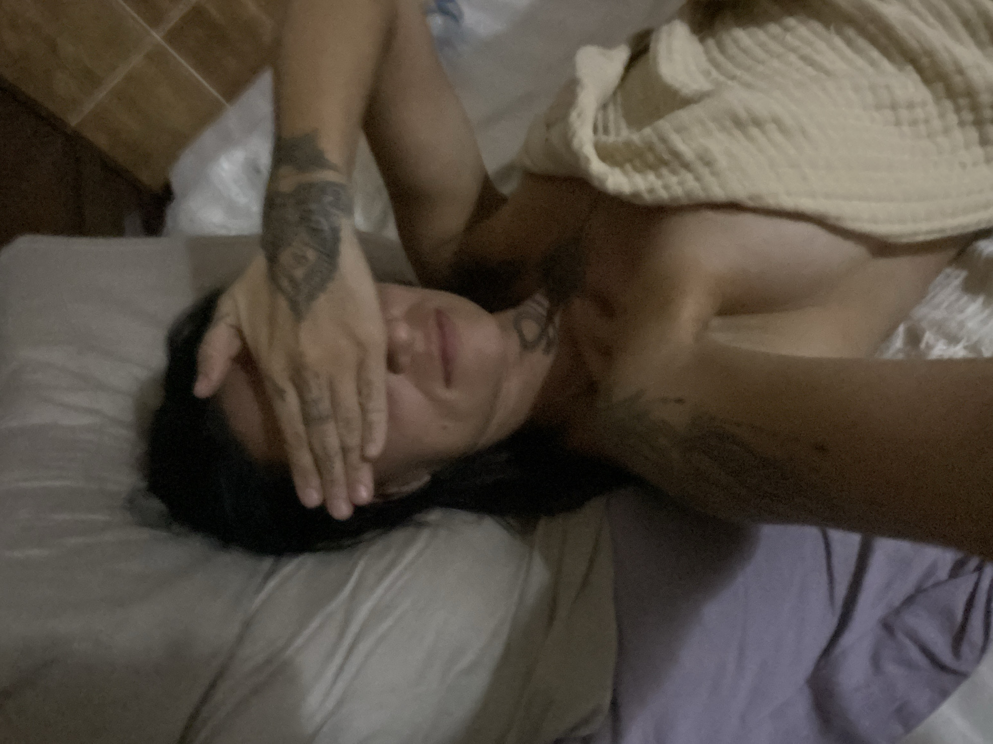 The scene is located in Mũi Né, Vietnam. An individual with long dark hair is lying on their back in a bed. Their left hand is raised, covering their eyes and upper face, with fingers spread slightly. A slight smile is visible on the lower half of their face, below the covering hand. Extensive tattoo work is visible on both arms, particularly on the left forearm and bicep, and on the right bicep and shoulder. A small tattoo is also present on the individual's neck/collarbone area. The individual's upper torso and chest are partially covered by a light-colored, textured blanket or towel, appearing to be a waffle-knit pattern. The bed features light-colored pillows and bedding, with a purple sheet visible towards the lower left. In the upper right background, a tiled floor with light brown square tiles and grout lines is visible, suggesting an indoor setting. The overall lighting is dim.