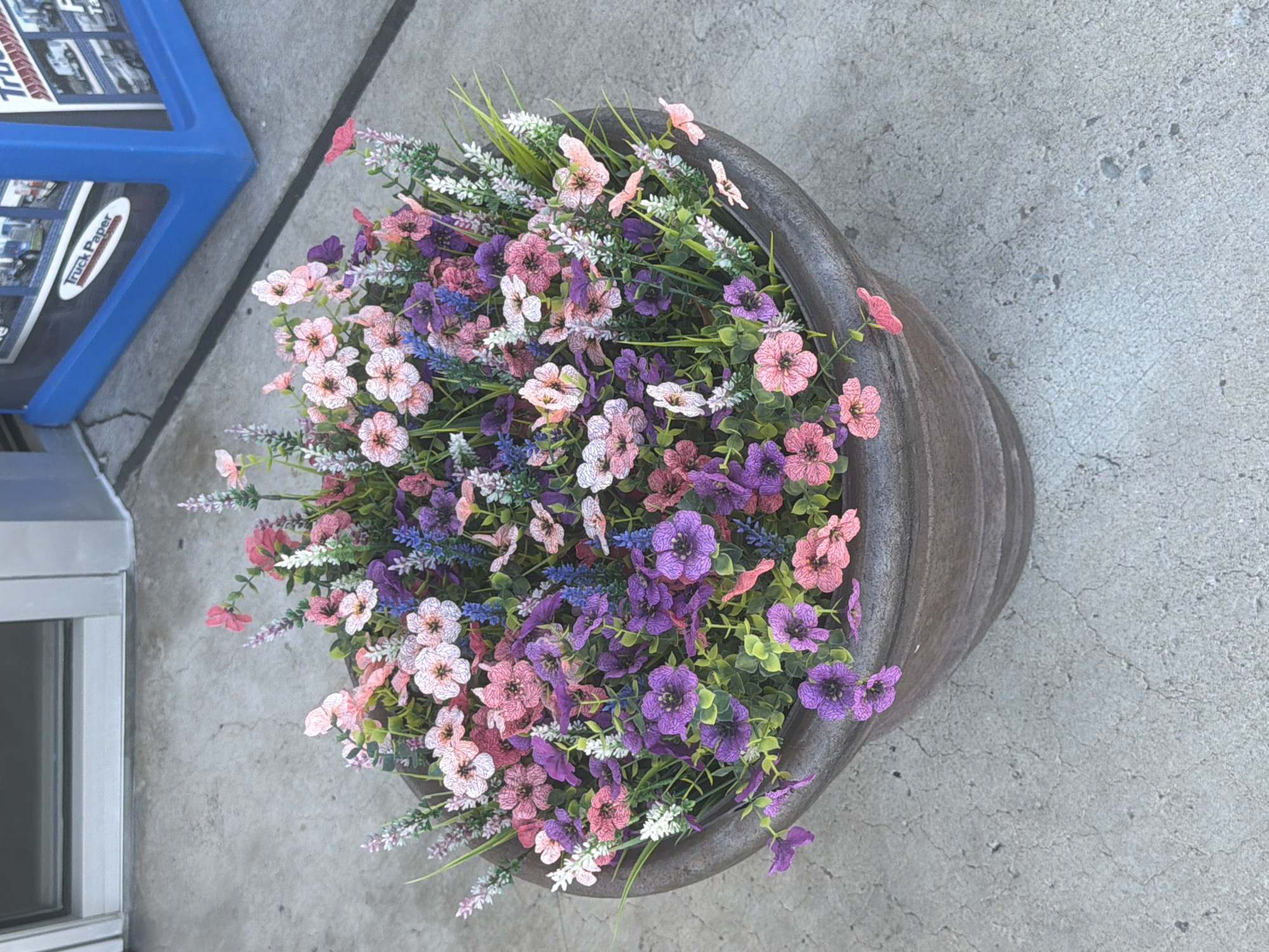 A large, round, dark brown planter filled with artificial flowers in various shades of pink, purple, and white sits on a gray concrete surface. The flowers are densely packed and include delicate, star-shaped blooms with darker centers, as well as spike-like clusters of lavender and tiny white flowers. Green foliage is interspersed throughout. In the background, a light-colored building with a horizontal window is visible, along with a blue stand displaying promotional material, possibly for a trucking publication, with the words "Truck Paper" clearly legible. The concrete surface shows a cracked texture and a dark line running across it. The scene appears to be outdoors, likely in front of a business. The location is the city of Lebanon, United States of America.