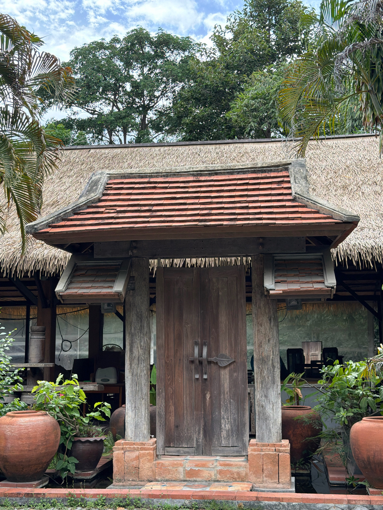 This is a full, eye-level outdoor shot of a wooden double door entrance to a building with a thatched roof and a red tile overhang. The entrance is flanked by thick wooden posts and a short brick platform. Two large terracotta pots with plants are visible to the left and right of the entrance. The background shows the interior of the building with furniture and some windows. The sky above is cloudy.