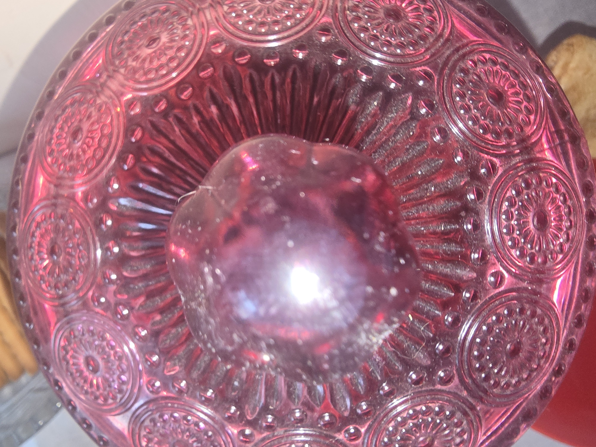 This is a close-up, top-down shot of a red glass bowl with intricate circular embossed patterns and a spherical object, possibly a plum or berry, resting inside. The object is dark red and reflects a bright light source. The bowl itself has a textured surface with raised circular designs featuring floral or sunburst motifs, separated by rows of small raised dots. The lighting creates strong highlights and shadows, giving the glass a glossy appearance. The background is blurred, showing hints of other objects, including what appears to be stacked bread or pastry. There is no indication of the location being Ulan-Ude, Russia; the image provides no geographical context.
