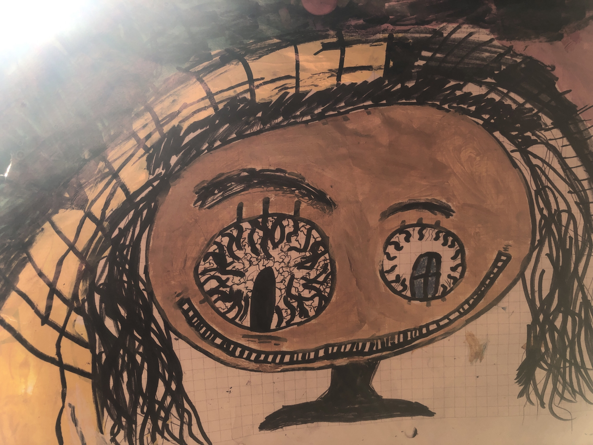 The image is a drawing of a stylized face with large eyes and a wide mouth. The face is painted in shades of brown, with black lines for hair and eyebrows. The eyes are intricate, with one eye containing what appears to be a tree-like structure, and the other containing a small window. The mouth is a wide, toothy grin. The background is a wash of yellow and brown with some darker abstract shapes. The drawing appears to be on grid paper. The lighting suggests a bright light source from the upper left, casting a glare across the image. There is no discernible location in a city of Unknown, Unknown.