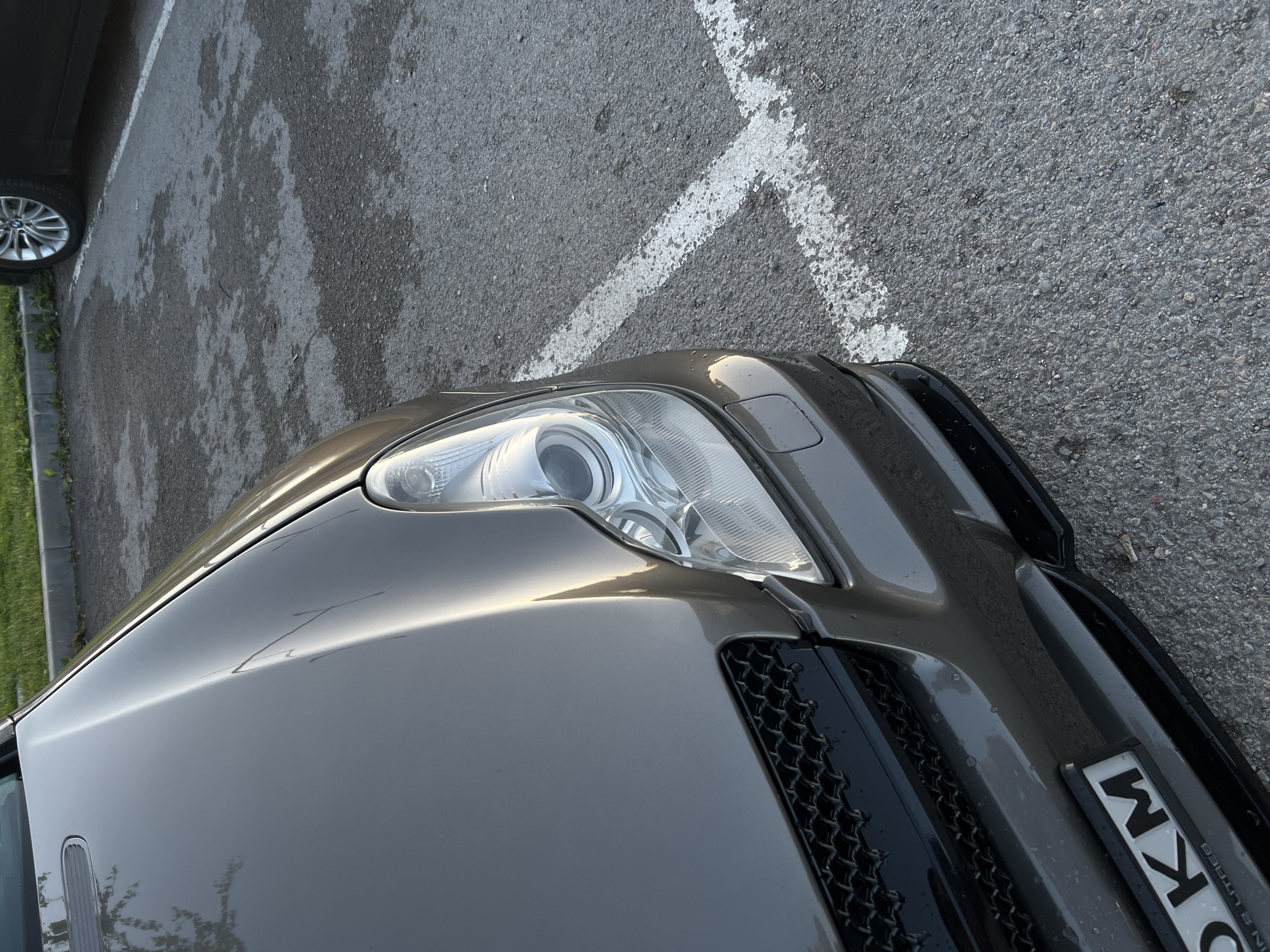 The image displays the front-right portion of a dark grey or brown car parked on textured asphalt pavement. The car features a sleek, modern design with a prominent headlight assembly, housing multiple circular and rectangular lighting elements. A black, honeycomb-style grille is visible towards the front of the vehicle. Reflections of trees and possibly the sky are cast upon the car's reflective hood and body panels.
To the left of the main car, a silver alloy wheel from another vehicle is partially visible, suggesting an adjacent parking spot. The asphalt pavement shows irregular wet patches, indicating recent rainfall. A thick, white painted line, likely a parking space marker, traverses the asphalt in front of the car. A narrow strip of green grass and a concrete curb are visible along the far left edge of the frame.
The natural lighting suggests either early morning or late afternoon, creating a soft sheen on the car's surface. On the front bumper of the main car, a partially visible license plate displays the letters "KM" clearly, followed by "O IN 2 LITRES", suggesting an engine size designation like "5.0 IN 2 LITRES." No people or specific activities are depicted; the scene portrays a static outdoor parking environment.