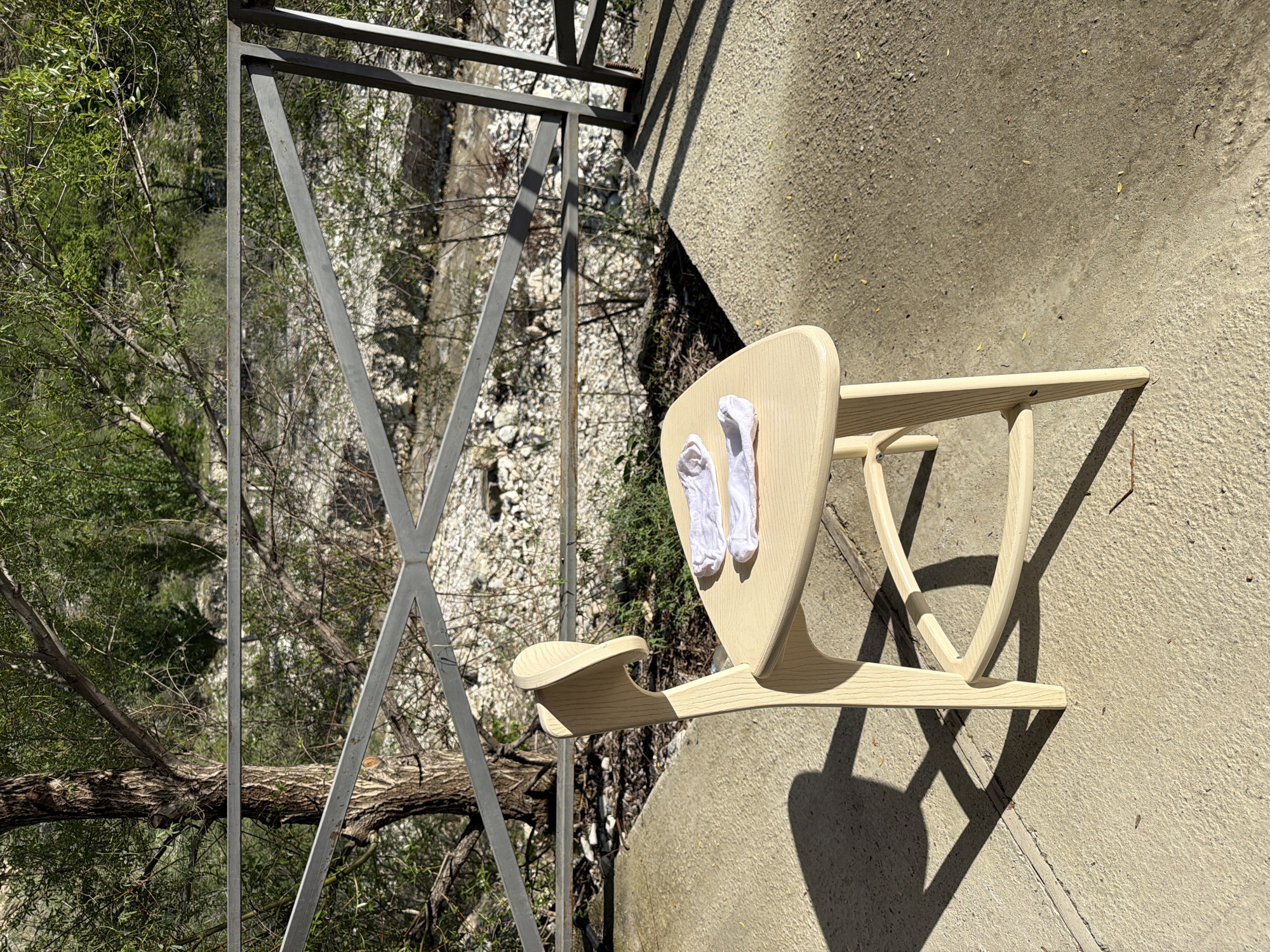 A slightly rotated eye-level shot reveals an outdoor scene, possibly a balcony overlooking trees and an unclear view beyond, potentially a riverbed or quarry due to the white stone visible. A light-colored wooden chair with two white socks placed on its backrest is centrally positioned. The chair casts a prominent shadow on the textured concrete surface of the balcony floor. A gray metal railing runs along the edge of the balcony. Beyond the railing, trees and a rocky area are visible. The city or town location is unknown, and the lighting suggests a sunny day.
